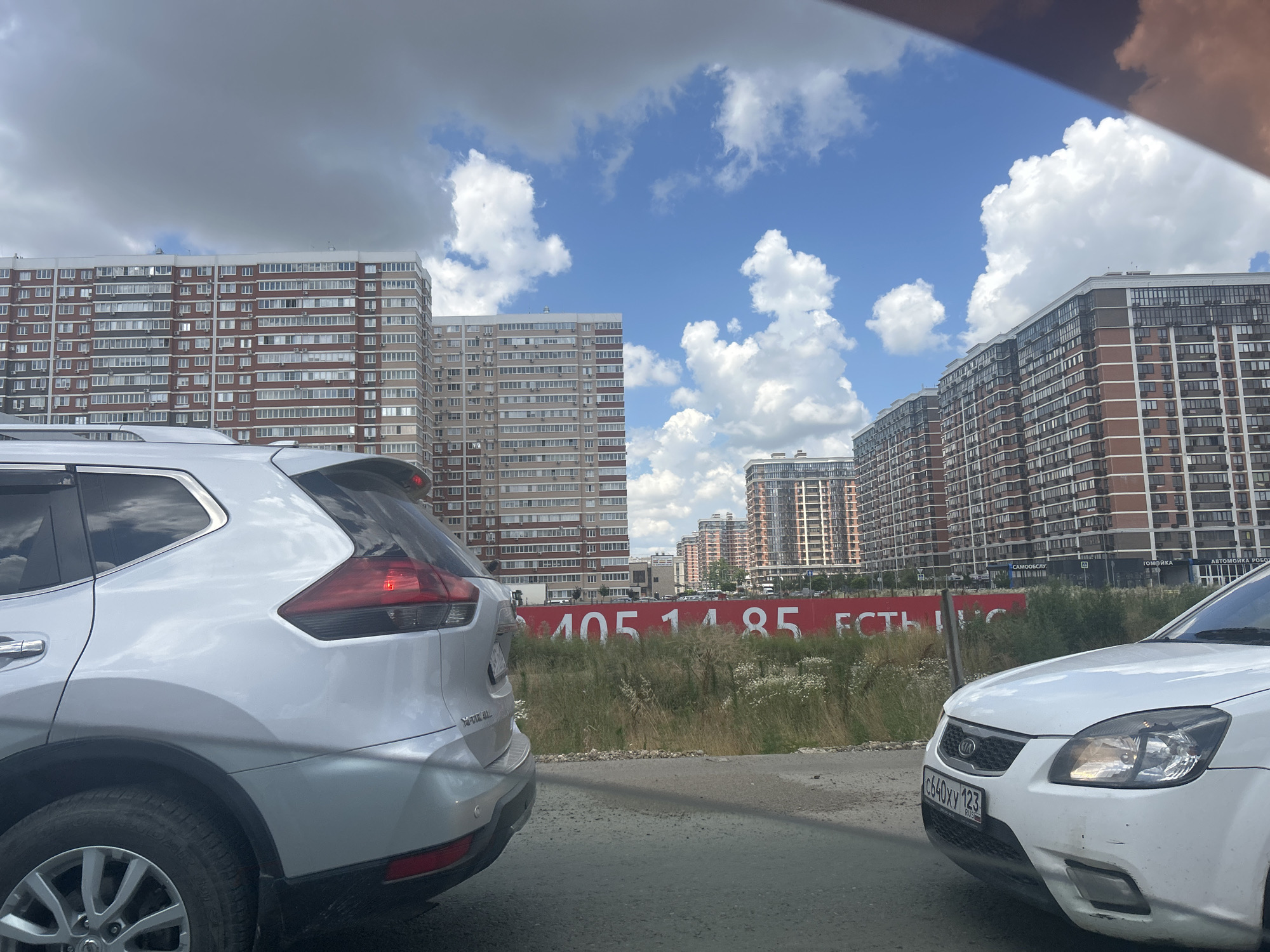 The image depicts a street scene in Orlovskii, Russia, with several tall apartment buildings dominating the background. The buildings are a mix of modern architecture with brick and glass facades. In the foreground, two cars are visible. On the left, the rear of a silver SUV is prominent, showing its taillight and part of its wheel. On the right, the front of a white Kia is visible, with its license plate displaying "c640XY 123" and the "RUS" indicator. A red banner stretches across the middle ground, adorned with white text that appears to be a series of numbers and partial words. The setting is outdoors, under a partly cloudy sky with significant fluffy white clouds against a bright blue backdrop. The time of day appears to be daytime, possibly late morning or afternoon, given the illumination. The environment suggests an urban or suburban residential area. There are no people clearly visible in the image.