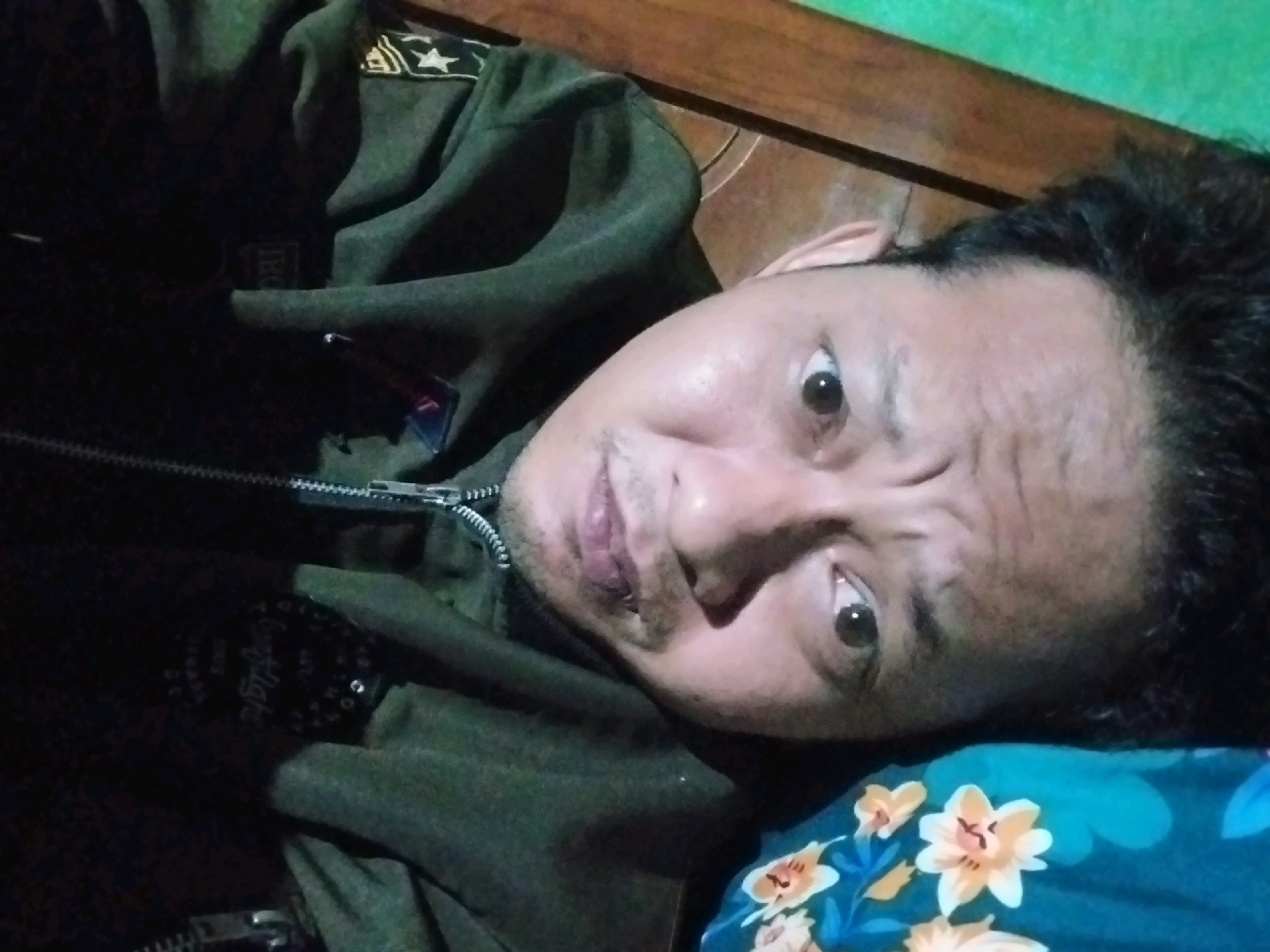 The image is a close-up, high-angle, self-taken photograph of a person lying down. The person appears to be male, with dark hair and brown eyes. He is wearing a dark green jacket with zippers and patches on the shoulder. The background consists of a wooden surface and a green fabric. The lighting is dim and appears to be artificial. There are no visible indications of the location in Campur, Indonesia, or any specific actions or interactions. The image is slightly blurry, suggesting it was taken with a hand-held device at a close range.