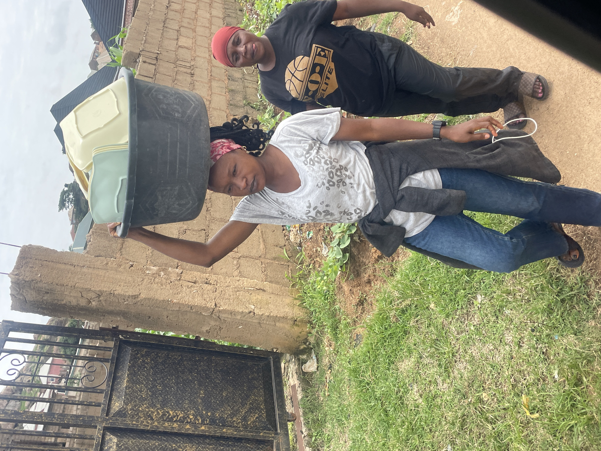 In Barakin Lerawi, Nigeria, two women walk on a dirt path beside a grassy area. The woman in the foreground, wearing a grey t-shirt and blue jeans, carries a large black tub filled with light-colored square dishes on her head. She has a red bandana tied around her head, and a dark grey sweater is tied around her waist. To her right, the second woman, wearing a black t-shirt with a basketball graphic and black pants, walks behind her. She has a red headscarf and is looking towards the camera. A metal gate with decorative scrollwork is visible to the left of the frame, and a partially constructed brick wall runs along the right side. The sky is overcast.
