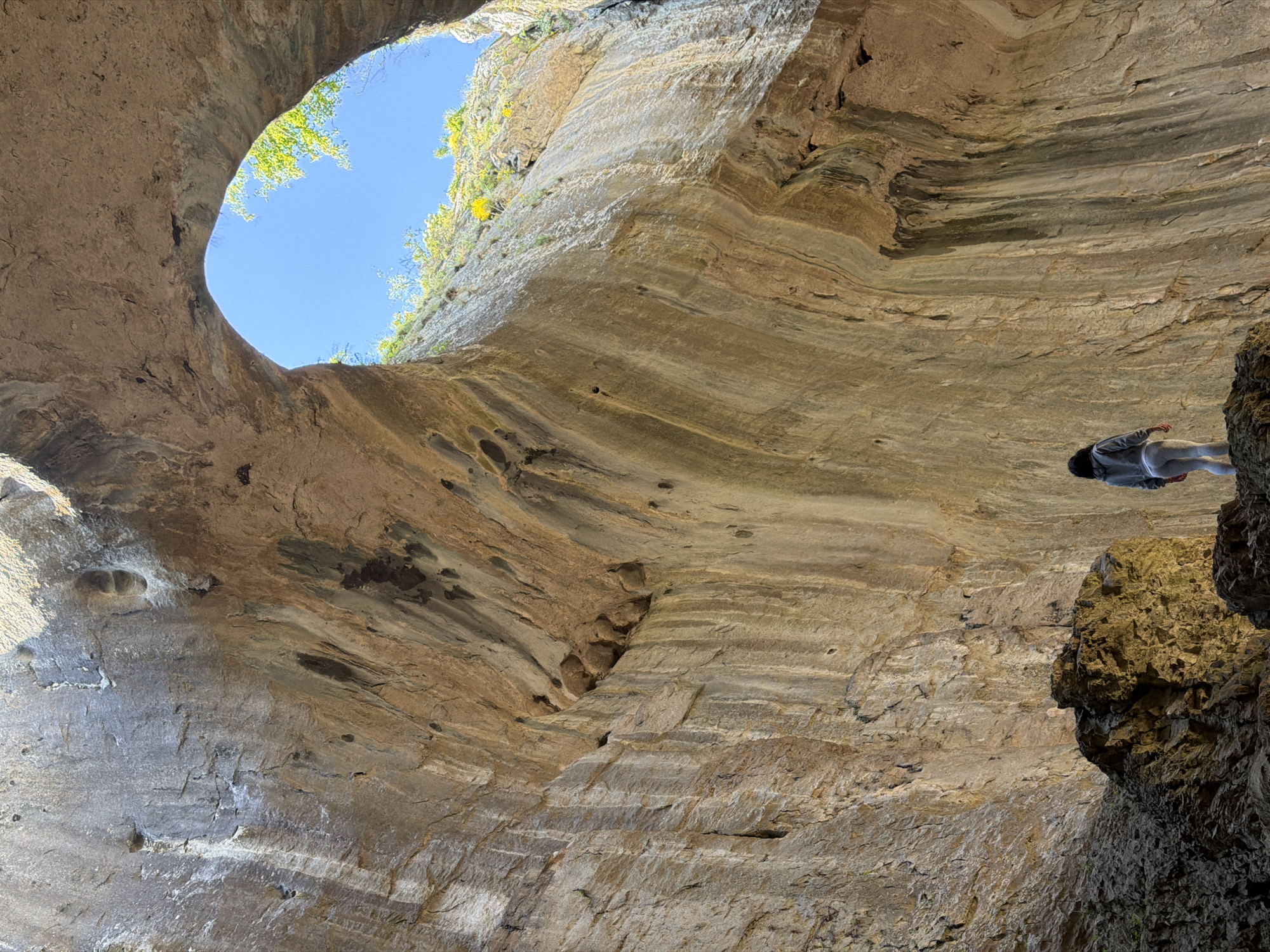 A low-angle shot captures the interior of a cavern. Layered rock formations in shades of beige and brown form the walls and ceiling. A circular opening reveals a bright blue sky and sparse green vegetation. A single person is visible on the right, precariously perched on a rock ledge, wearing light clothing and facing away from the viewer. The location is Karlukovo, Bulgaria, judging by the cavern's formation and vegetation.