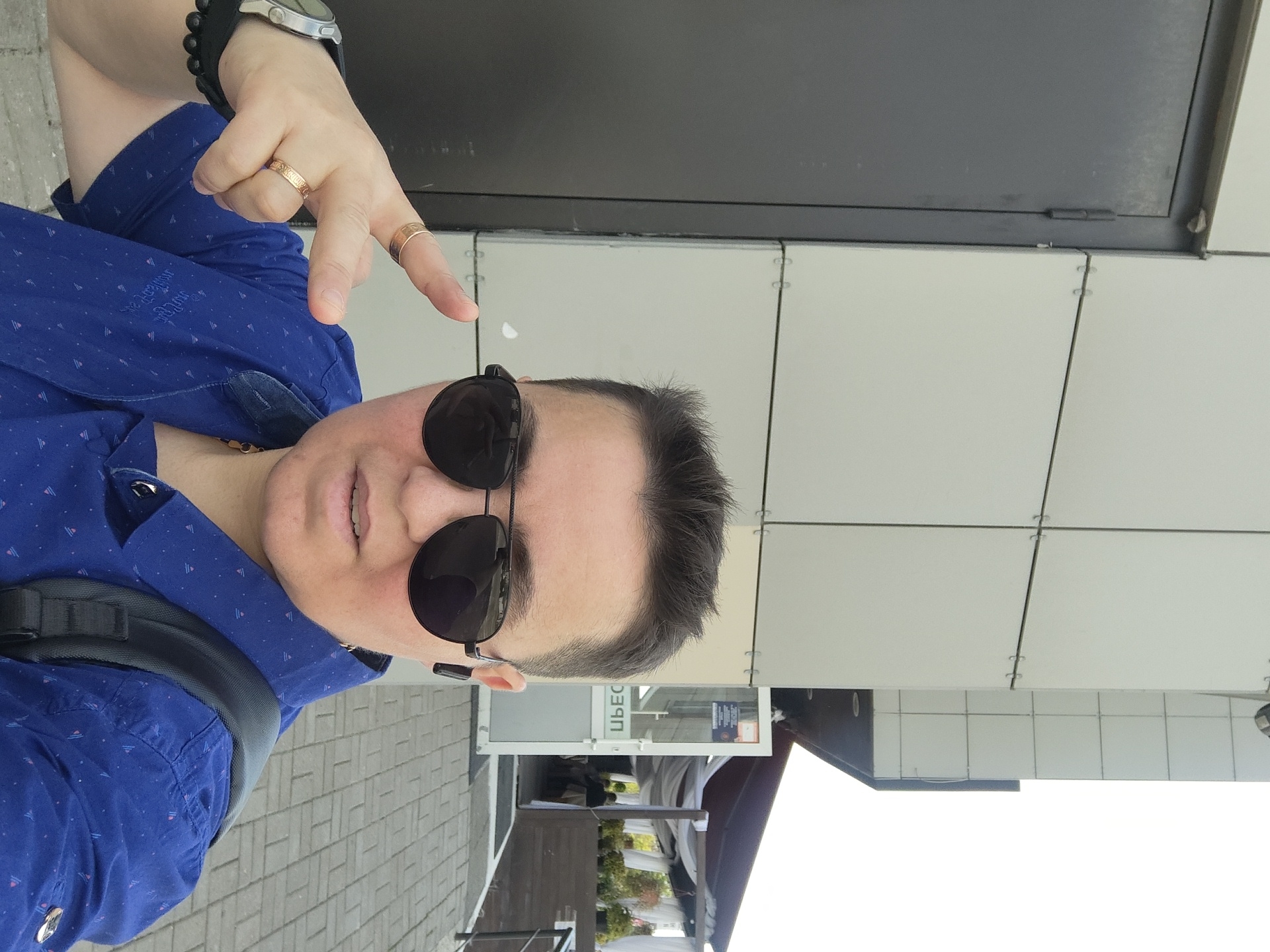 A person stands in front of a building with a gray metal door and a tiled facade. The person is wearing sunglasses, a blue patterned shirt, and has rings on their fingers. A backpack is visible on their shoulder. In the background, there is an entrance to a building with a canopy and outdoor seating, suggesting a cafe or restaurant. The ground is paved with brick-like stones. The scene appears to be an outdoor urban setting, possibly in the city of Zhukovsky, Russia.