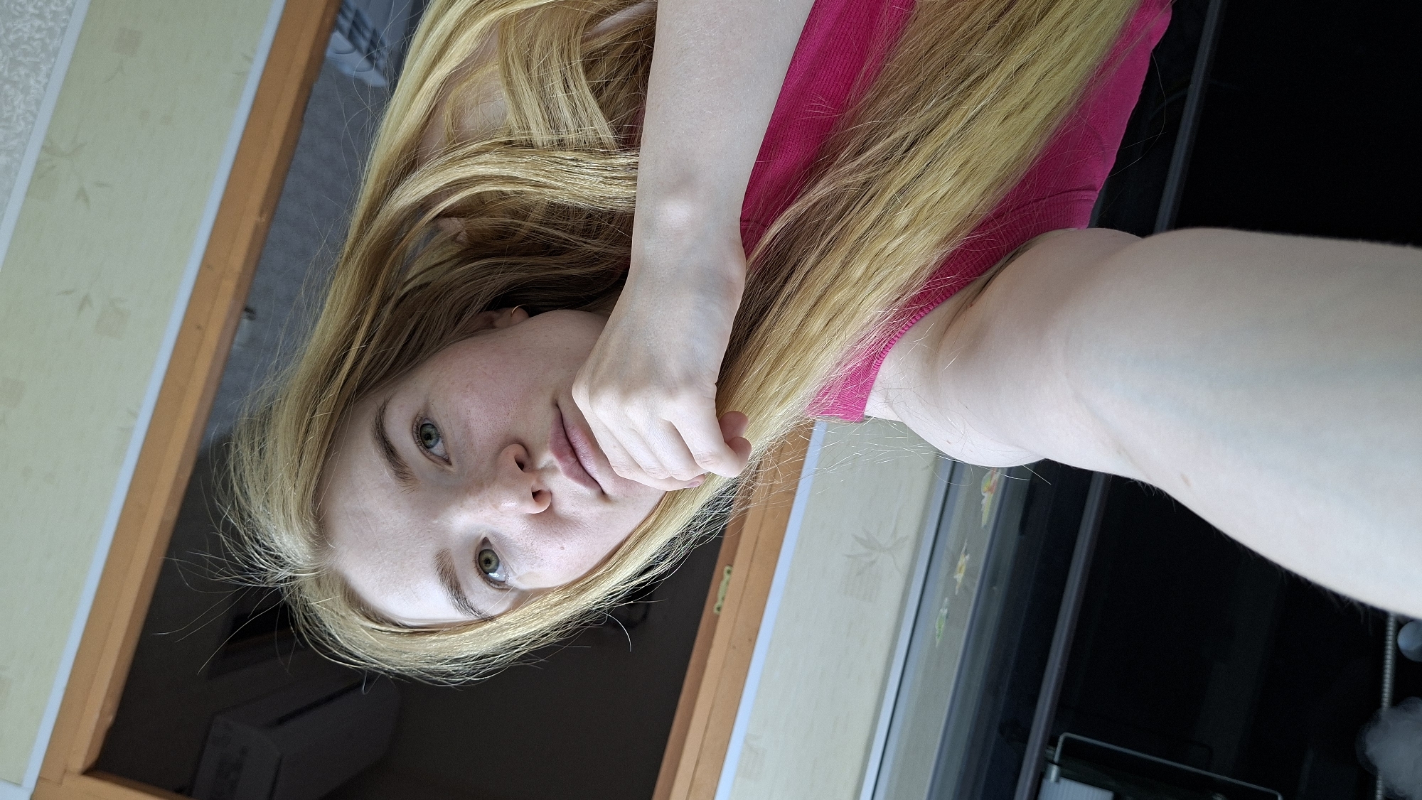 This media file is a portrait of a young woman, captured in a selfie-like shot from an overhead perspective, with the image rotated 90 degrees counter-clockwise. The woman has long, straight, light blonde hair that flows around her head and across her shoulders. She possesses fair skin, distinct green eyes, and light eyebrows, with some faint freckles visible on her face. She is wearing a bright fuchsia or hot pink sleeveless top or dress. Her right hand is positioned near her mouth or chin, creating a casual or contemplative pose.
The background indicates an indoor setting, likely by a window or glass door. To her left, a wall with light-colored, possibly beige, wallpaper featuring a subtle floral or leaf pattern is visible. A wooden frame, possibly part of a window or doorway, runs vertically along this wall. To her right, a dark, somewhat reflective area suggests an exterior view or another room through a pane of glass. A blurry white object, possibly an air conditioning unit or a fixture, is seen in the bottom left background. The natural, bright lighting suggests the photo was taken during the daytime. There is no visible text in the image. The location context provided is Большой Букор, Russia.