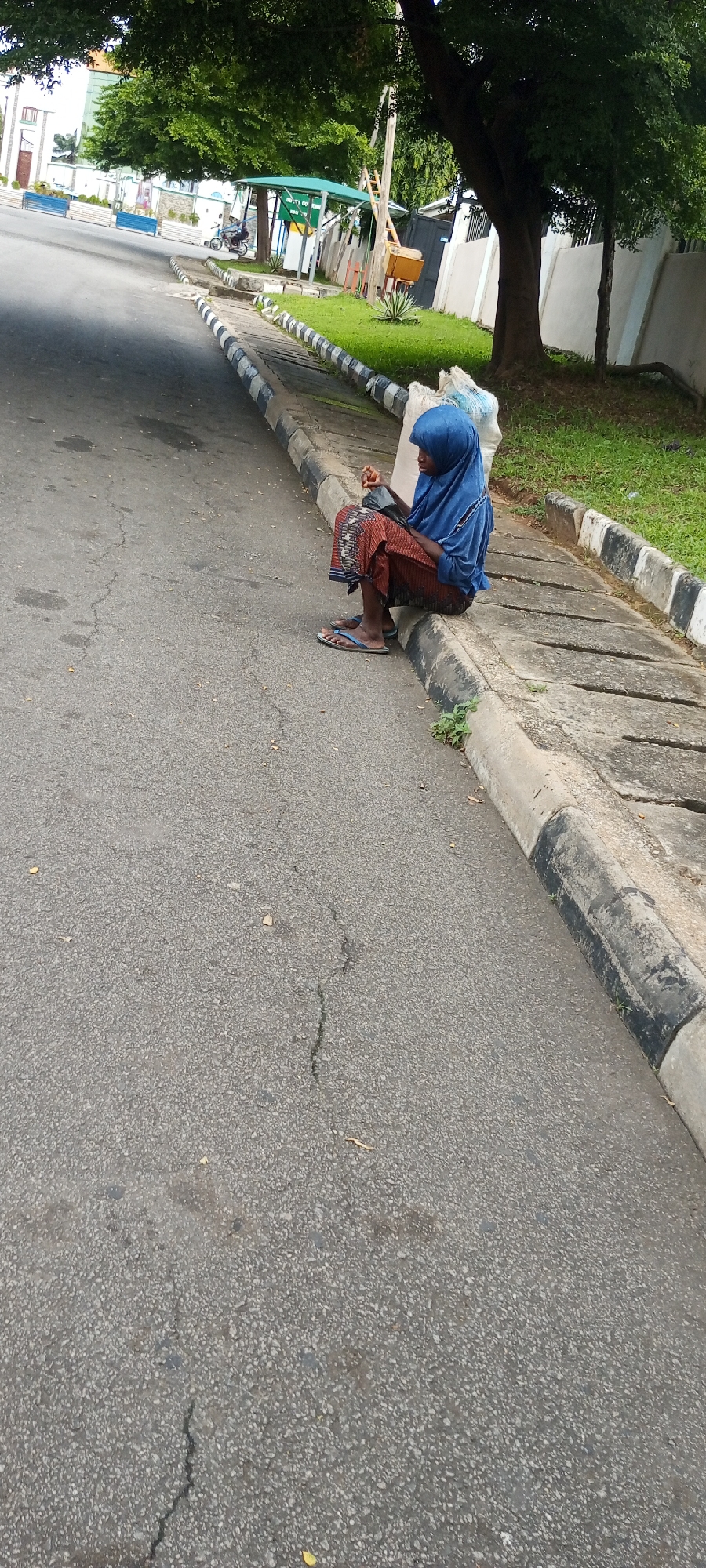 A person wearing a blue hijab and a patterned skirt sits on the edge of a road in Adankolo, Nigeria. The person is facing right, holding a dark object in their hands. Two white bags are behind them. The road is paved with asphalt and has a concrete curb with black and white markings. Green grass and trees are visible beyond the curb. In the background, there is a green shelter with a motorcycle parked nearby and a ladder leaning against it. A fence and buildings are also visible in the distance.