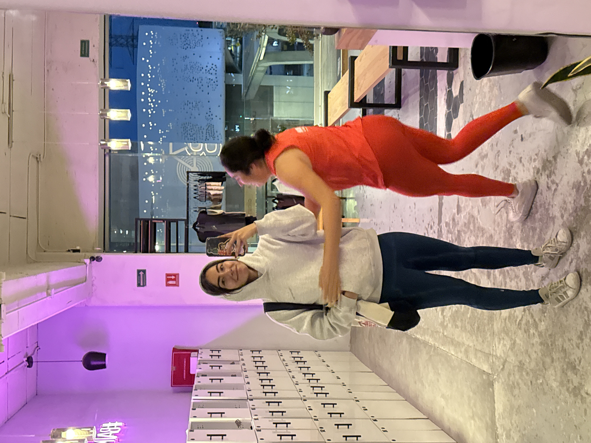 Two women are posing for a photo in what appears to be a gym or fitness studio. The woman in the foreground wears a grey hoodie and blue leggings. She is holding a phone and smiling at the camera. Behind her is a wall of lockers. The other woman, in the background, wears an orange tank top and matching leggings. She is in a plank position, suspended between a bench and the floor. Outside the window, an urban scene is visible, including a tall building with patterned lights. A green emergency exit sign and a red fire extinguisher symbol are visible on the wall. The photo suggests the location is likely in Puebla, Mexico, based on the visible building details.
