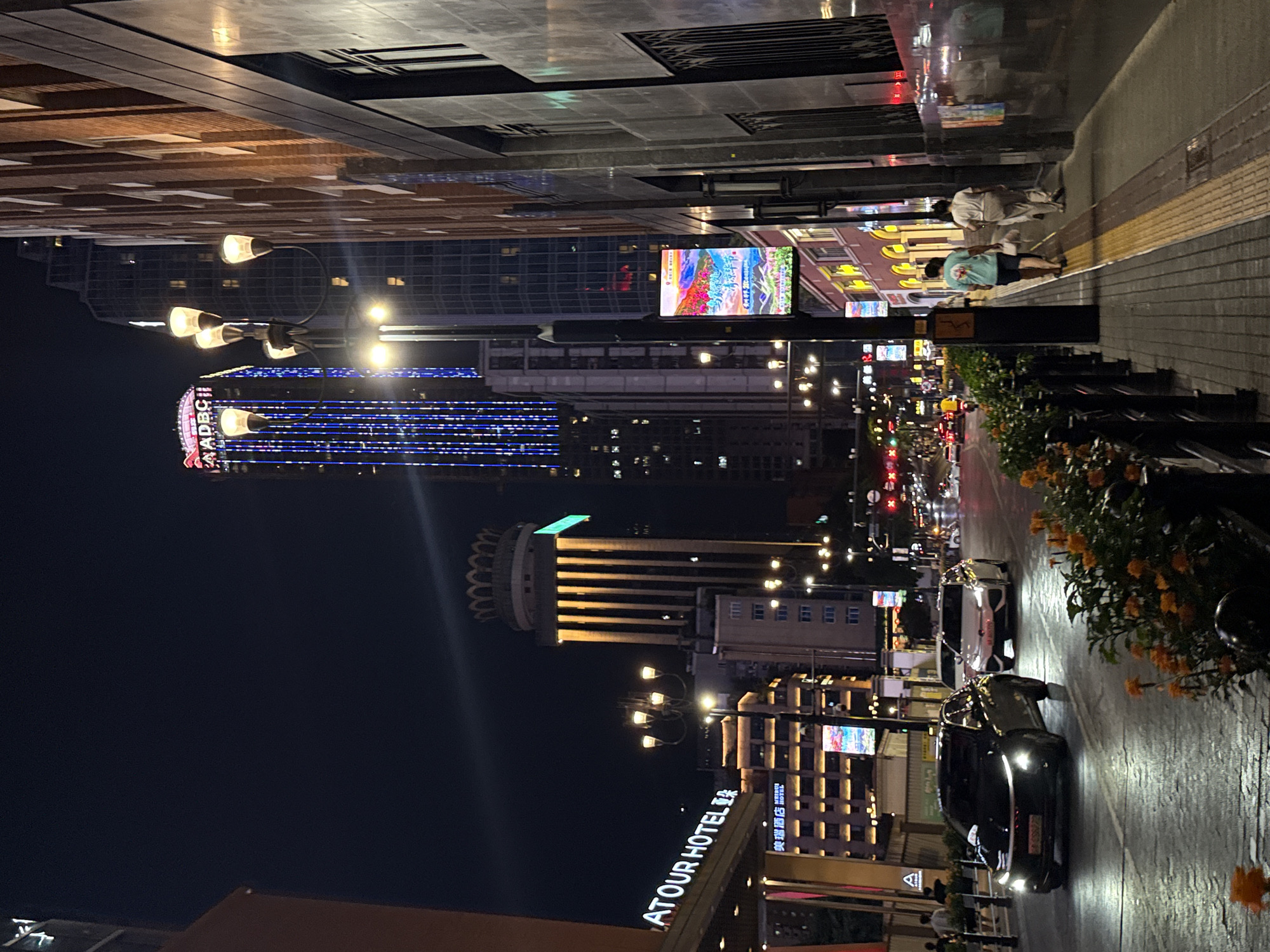 This is a night-time photograph of a street scene in the Yuzhong District of China. The street is lined with multi-story buildings, some of which are illuminated with colorful lights. A tall skyscraper with blue lights and the text "ADBC" at its peak is visible in the background. Several cars are driving on the wet street, reflecting the lights from the buildings. On the right side of the street, a sidewalk runs alongside the buildings, with two people walking away from the camera. Orange flowers are planted in planters along the edge of the sidewalk. A street lamp with multiple lights is also visible. The overall impression is of a busy urban environment at night.