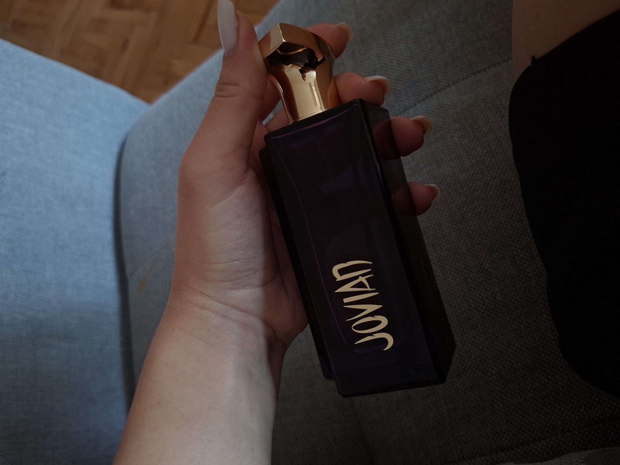 A hand holds a bottle of Jovian perfume in a dimly lit, indoor setting. The bottle is dark purple with a gold-colored cap and the brand name "JOVIAN" written in gold lettering down its side. The hand is fair-skinned, with long manicured nails. The background consists of a gray textured sofa and a hint of a wooden floor. The location in the city of Pleven, Bulgaria is not discernible from the image.