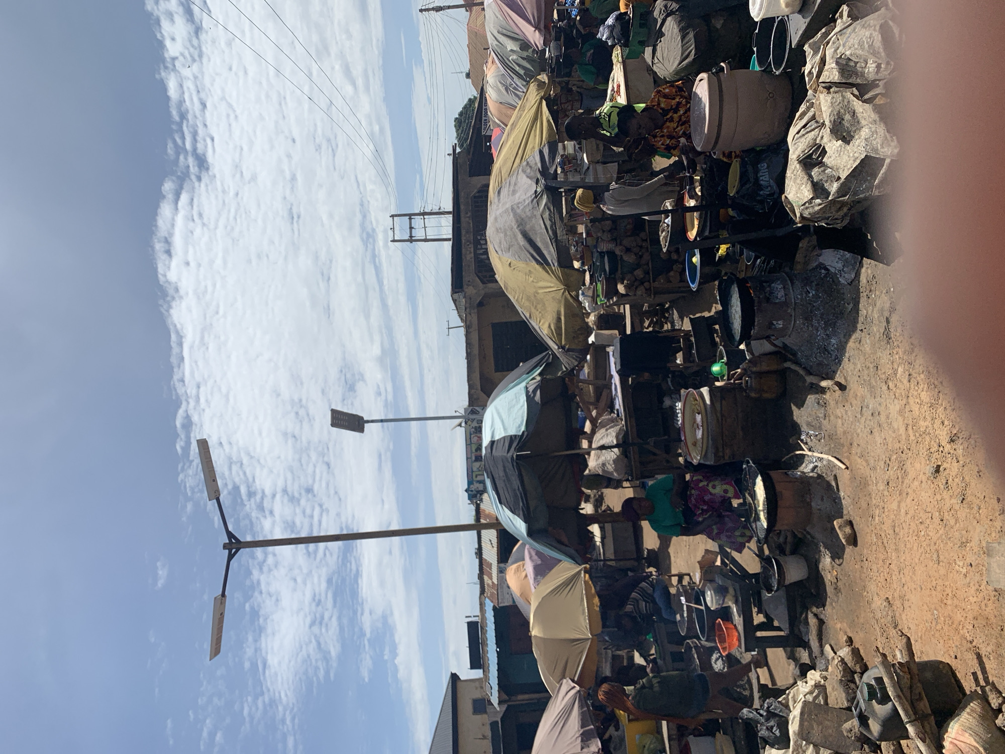 This image depicts an active outdoor market scene in Rafin Par, Nigeria, under a partially cloudy sky with bright daylight. The ground consists of dry, reddish-brown earth. In the foreground, a blurry human finger partially obscures the bottom right corner.
**Infrastructure and Environment:**
Two modern, solar-powered streetlights on tall poles are visible against the sky; one has a dual-arm fixture, and another single-arm fixture is to its right. Multiple power lines traverse the upper right quadrant, connecting to an electrical tower-like structure in the background. Buildings with varying roof types, including corrugated metal and flat, are present in the middle and background, suggesting a mixed-use or commercial area.
**Market Setup and Objects:**
Numerous large market umbrellas of various colors, including brown, gold, black and light blue, and olive green, are pitched to provide shade over vendor stalls. Beneath these umbrellas, goods are displayed on wooden tables and directly on the ground. Visible items include piles of produce (e.g., yams or similar tubers), plastic basins, metal pots, and various containers. Several large, light-colored ceramic or plastic containers are also present.
**People and Activities:**
Approximately ten to fifteen individuals are discernible within the frame, primarily women. People are engaged in various market activities:
* A woman in a green shirt and patterned skirt sits on a low stool next to a stall with a large white basin, possibly tending to her goods.
* Another woman in a yellow headwrap and light-colored top stands near a stall.
* In the foreground, two circular charcoal stoves, constructed from concrete or clay, have pots placed on them. One pot contains cooking oil or food being fried, indicative of food preparation or sales.
* Other individuals are seen standing or sitting, appearing to be vendors or shoppers.
* A black jerrycan, some wooden sticks, and miscellaneous debris are scattered on the ground near the cooking area.