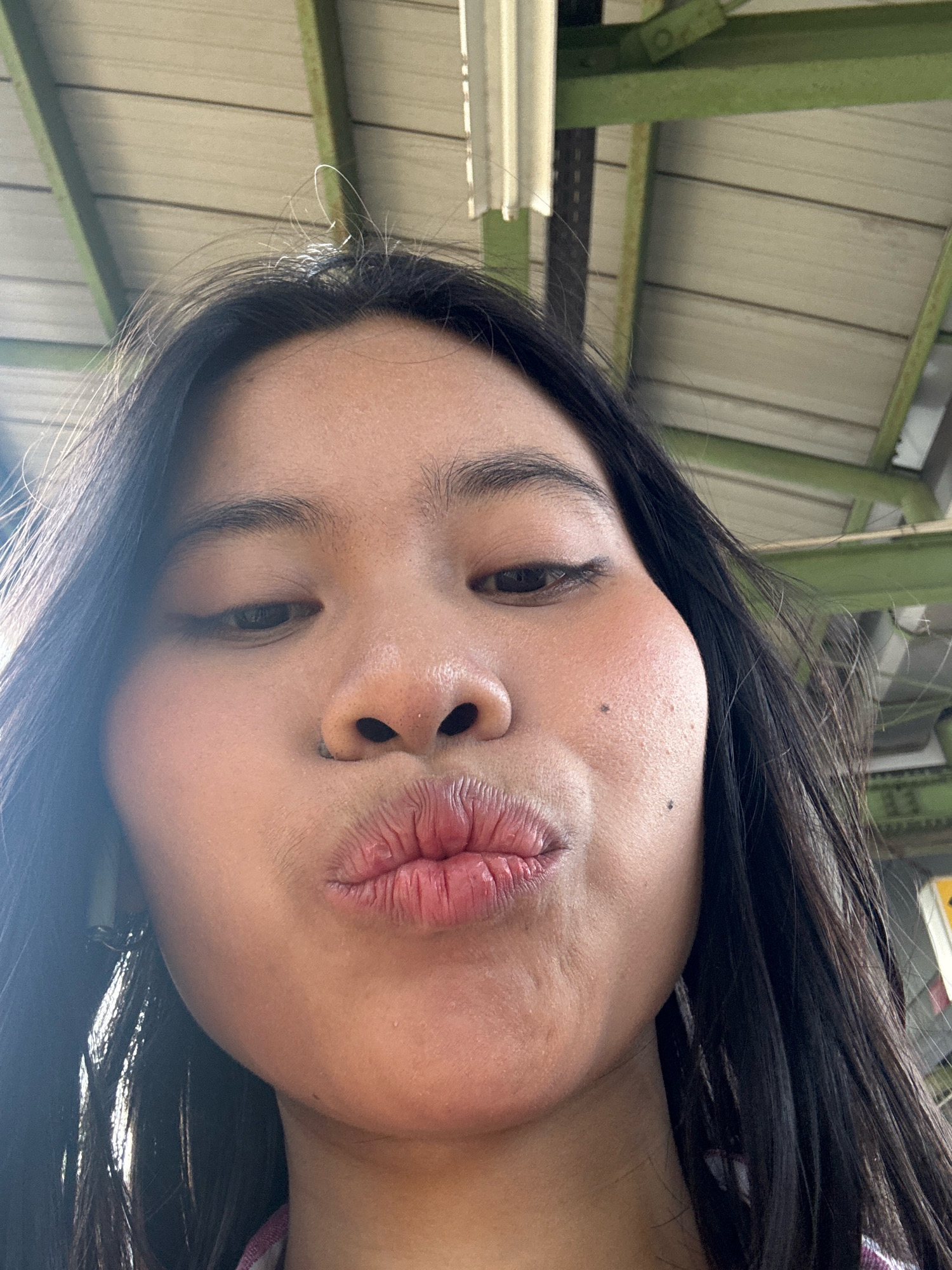 A close-up, selfie-style, waist-up shot features a young Asian female with long, dark hair, pouting her lips. She is wearing a pink and white plaid top. The background consists of a corrugated metal ceiling with green beams, indicative of an outdoor covered area, possibly a bus stop or market shelter, in East Jakarta, Indonesia. The lighting suggests it is daytime.