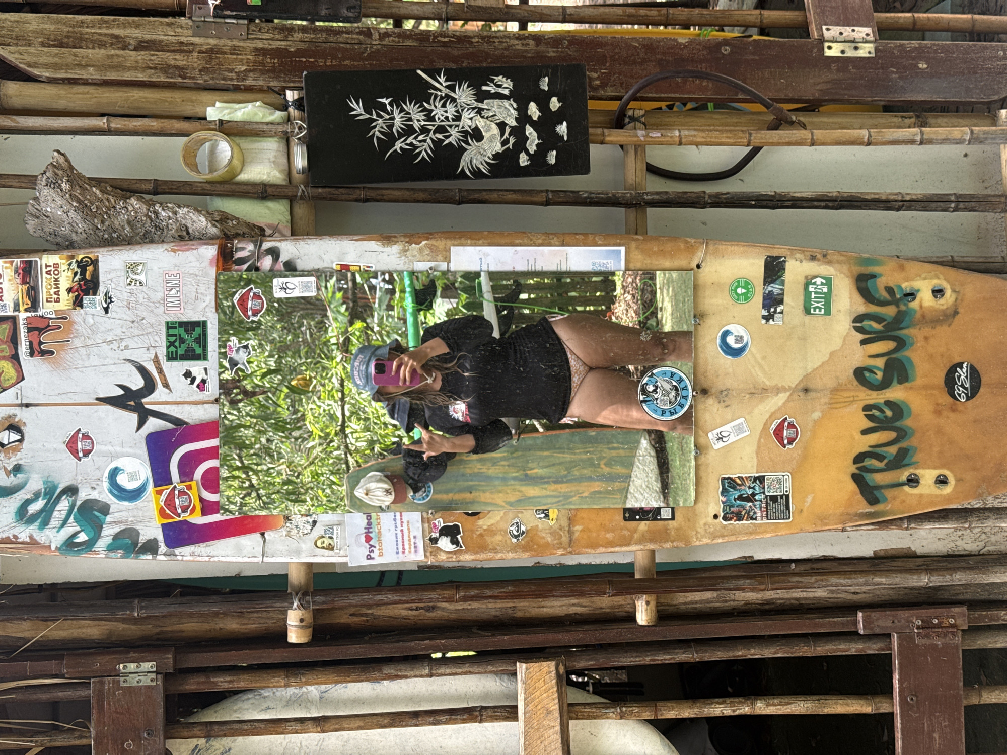 The image depicts a surf shop or surf school in Cam Lam, Vietnam. A woman, wearing a blue hat, black long-sleeve shirt, and patterned bikini bottoms, is taking a mirror selfie in front of a surf shop. She is holding her phone and making a "rock on" hand gesture. The surf shop displays a collection of surfboards adorned with various stickers and graffiti. The environment appears to be outdoors, with wooden structures and lush greenery visible in the background.