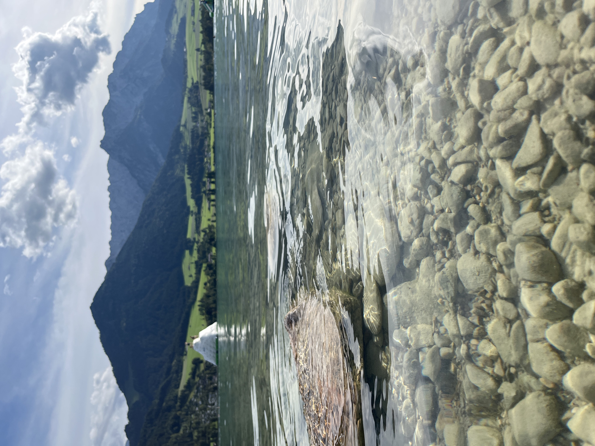 This is a close-up, low-angle shot of clear, shallow lake water revealing a rocky bottom. In the foreground, a submerged rock is partially visible, breaking the water's surface. The water exhibits ripples and reflections. In the mid-ground, the lake extends towards a distant shore lined with trees and a village. An inflatable water structure, possibly a slide or obstacle course, is present on the water near the shore. The background features rolling green hills and a majestic mountain range under a partly cloudy blue sky. The scene is located at Walchsee, Austria, a picturesque mountain lake known for its recreational activities.