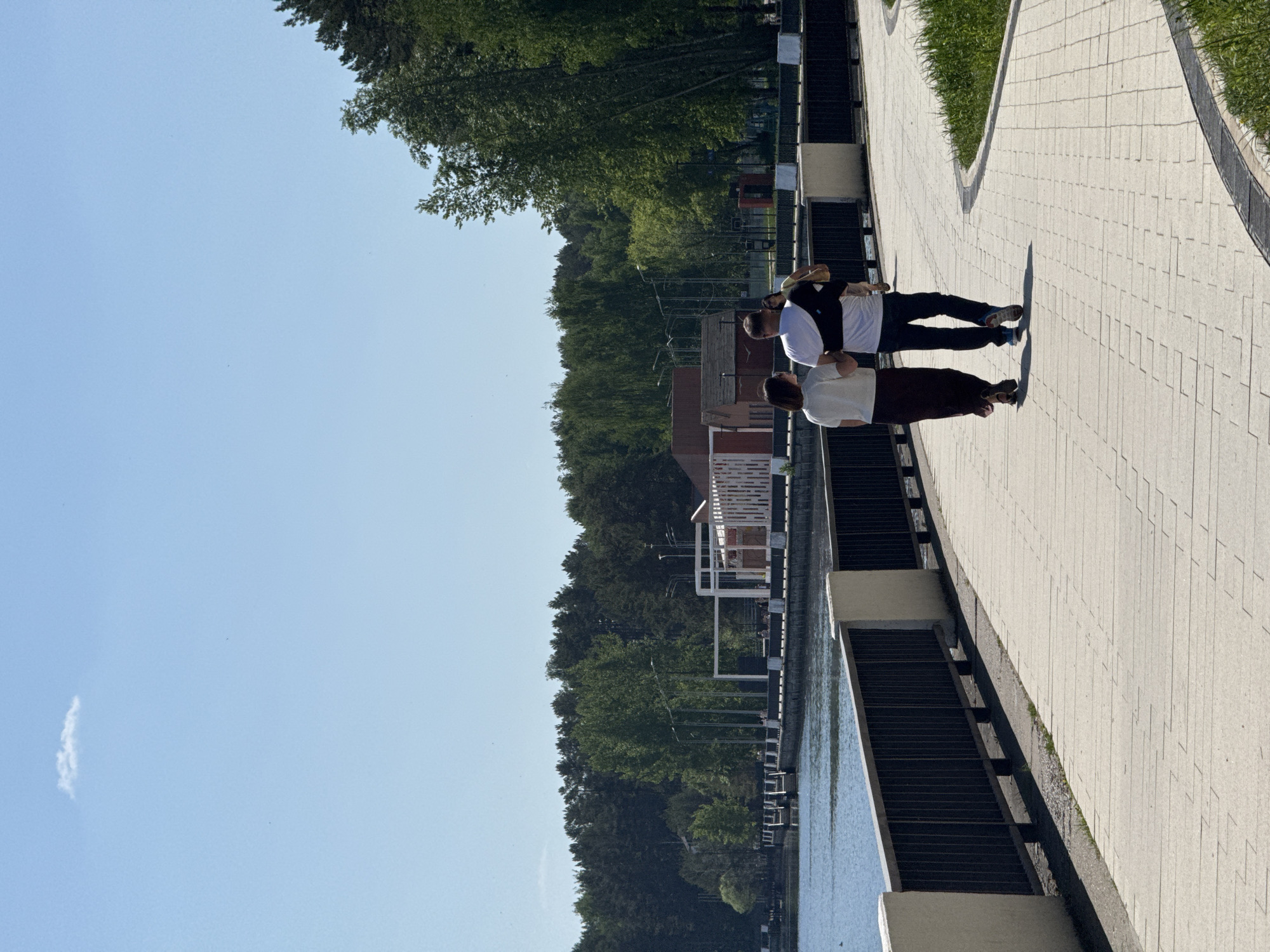 Two people are walking along a paved path in a park setting. The path is made of light-colored rectangular stones. One person is wearing a white shirt and dark pants, while the other is wearing a white shirt with a dark vest and dark pants. The path is bordered by a low wall on one side and grass and trees on the other. There is water visible beyond the wall. The sky is blue with some light wispy clouds. A building is visible through the trees in the background.