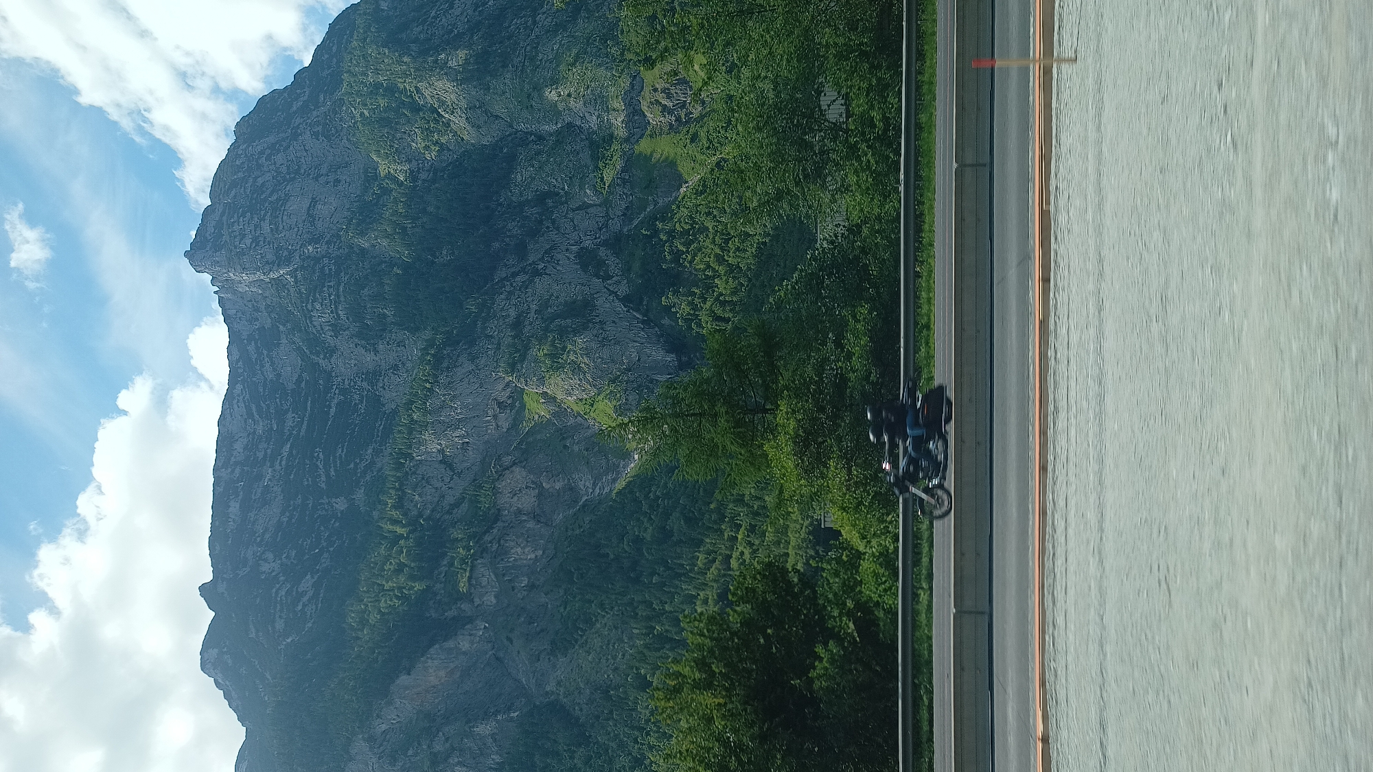 This image captures a mountainous landscape and a road in Golling an der Salzach, Austria. The upper two-thirds of the frame are dominated by a massive mountain range, characterized by steep, rugged grey rock faces interspersed with dense green coniferous forests. Lighter green vegetation patches are visible on some steeper mountain sections. Above the mountains, the sky is partly cloudy, with areas of clear blue visible between white cumulus clouds.
A multi-lane paved road runs horizontally across the mid-ground, separated by a grey metal guardrail and a concrete barrier. On the far side of this road, a dark-colored motorcycle is in motion, traveling from left to right. Two individuals, both wearing dark helmets, are riding the motorcycle. The immediate foreground consists of a light grey, textured surface, possibly gravel or an unfinished road. A slender wooden stake with a red tip is positioned vertically on the far right of this foreground. Motion blur is evident on the motorcycle and the immediate foreground, suggesting the photograph was taken from a moving vantage point.