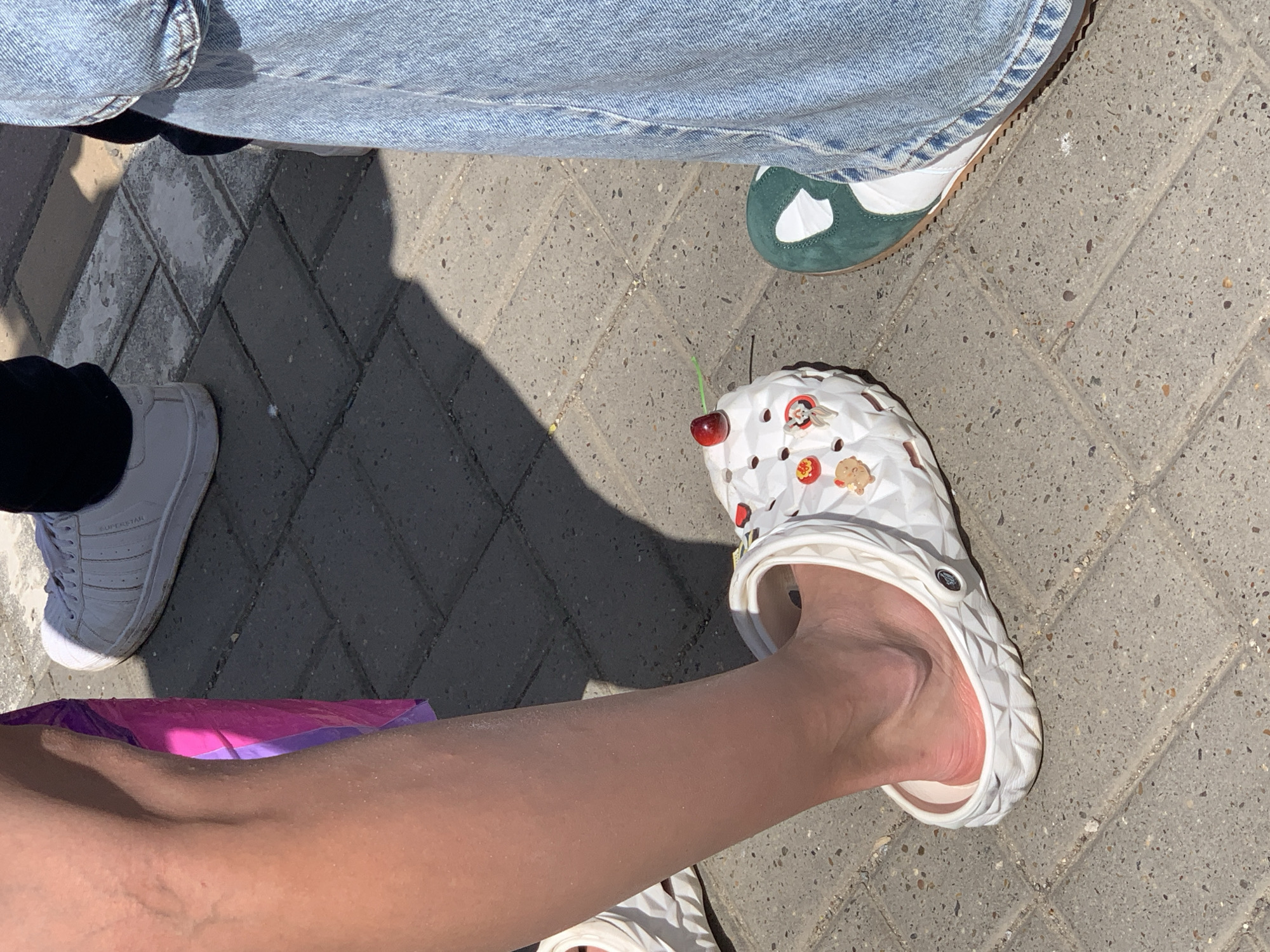 The image shows a close-up of three pairs of shoes on a paved surface, likely a sidewalk in a city. The shoes are:
1. A pair of light-wash denim jeans with a shoe peeking out from under the hem, showing a white and green sneaker.
2. A white sneaker with black trim.
3. A pair of white Crocs with various decorative charms. A fresh cherry with its stem is lodged in one of the holes.
The paved surface is made of grey bricks in a herringbone pattern. There are shadows cast on the surface, suggesting a sunny day. The bottom of the frame features a leg wearing the Crocs. Part of a bright pink and purple bag is also visible. The location is not possible to determine from the image.