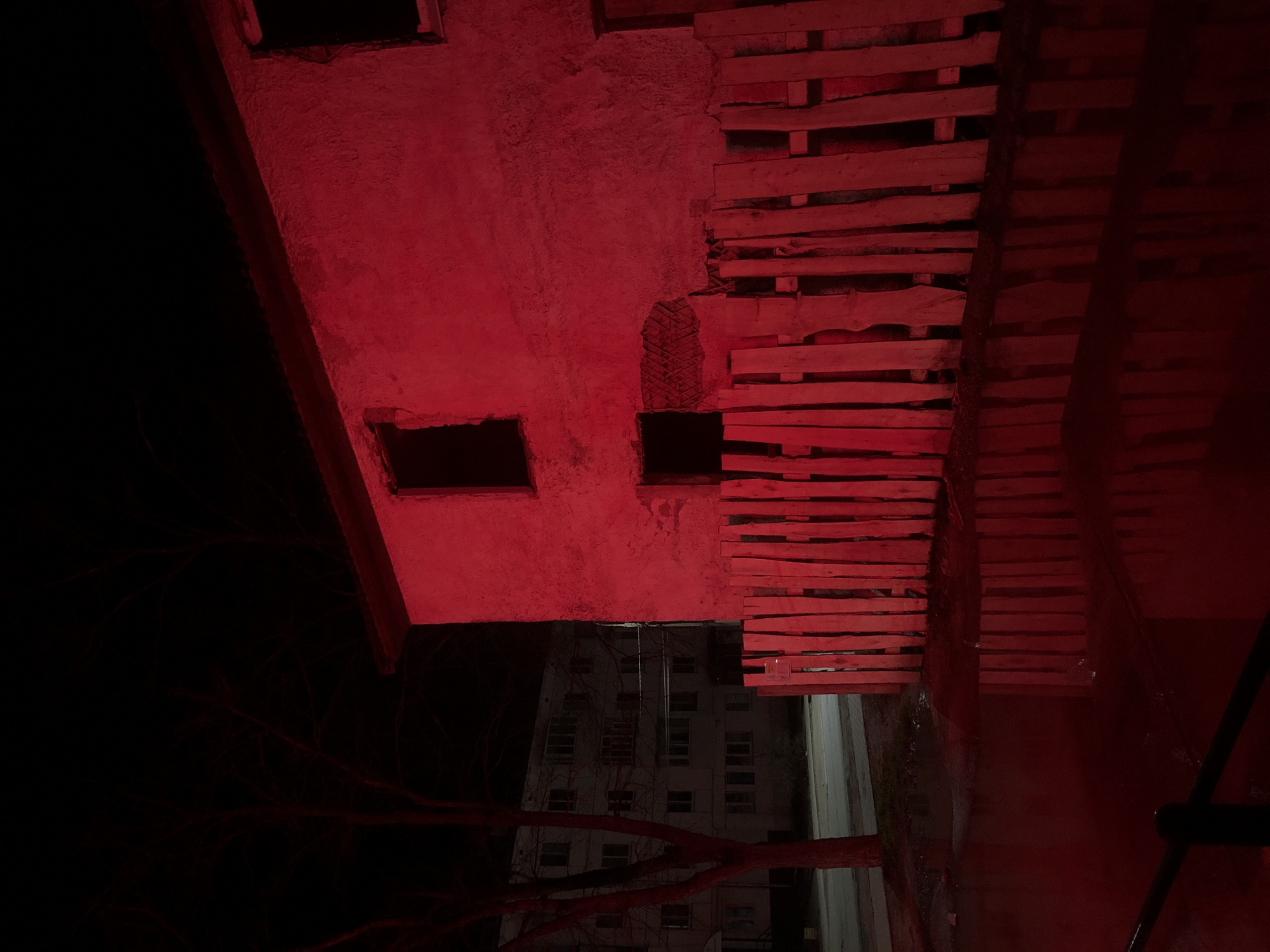The image shows the exterior of a weathered building at night in Chaikovsky, Russia. The building appears to be made of concrete or brick and has rectangular window openings. Adjacent to the building is a makeshift structure of stacked wooden planks. The scene is illuminated with a red light source, casting an intense red hue over the architecture and surrounding elements. In the background, a multi-story building with rows of windows is visible. The foreground is largely dark with a few bare tree branches. The overall impression is one of urban decay and makeshift construction under unusual, intense lighting conditions.