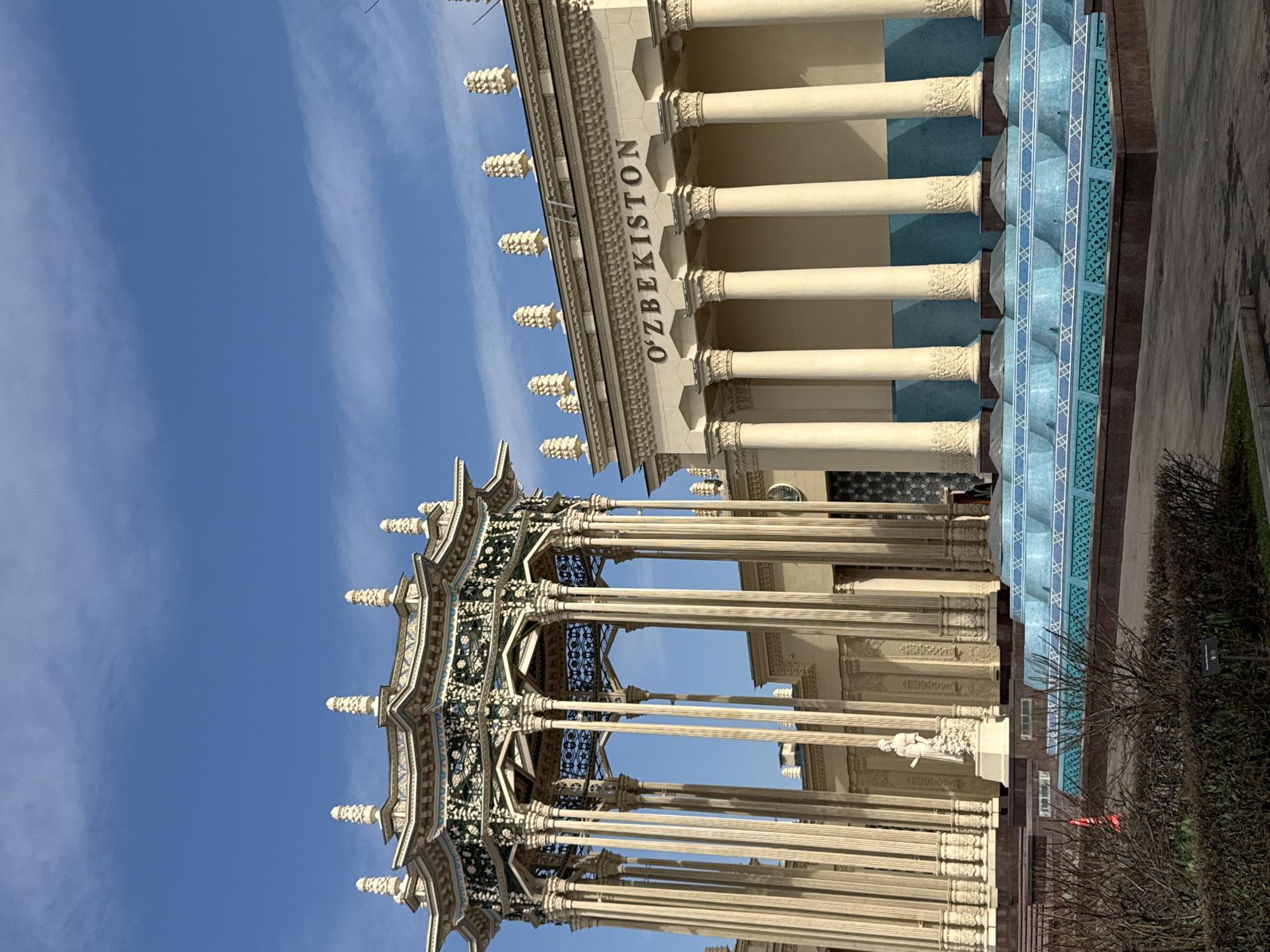 The image shows a building complex in Moscow, Russia, specifically the Uzbekistan Pavilion at the All-Russia Exhibition Centre (VDNKh). The main features include a two-story building with the name "O'ZBEKISTON" inscribed on its facade, supported by numerous columns and topped with decorative elements. Adjacent to it is an intricately designed, multi-tiered structure with an open-air pavilion, also supported by slender columns. The backdrop is a clear blue sky with wispy clouds. The foreground includes greenery, suggesting a park setting.