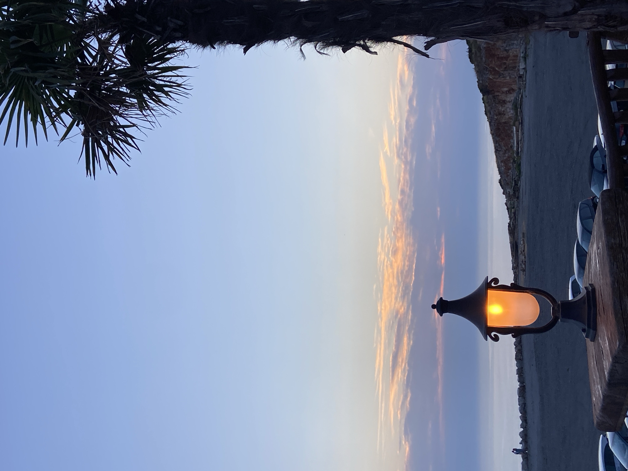 An outdoor scene in Kovačko Polje, Montenegro, features a lit lantern in the foreground, casting a warm orange glow against a twilight sky. A palm tree's fronds are silhouetted against the blue and pink hues of the sunset clouds. In the mid-ground, a body of water meets a rocky coastline, with a stone wall and a parking lot visible. Several cars are parked in the lot. The overall atmosphere is serene, with the natural beauty of the landscape illuminated by the artificial light of the lantern.