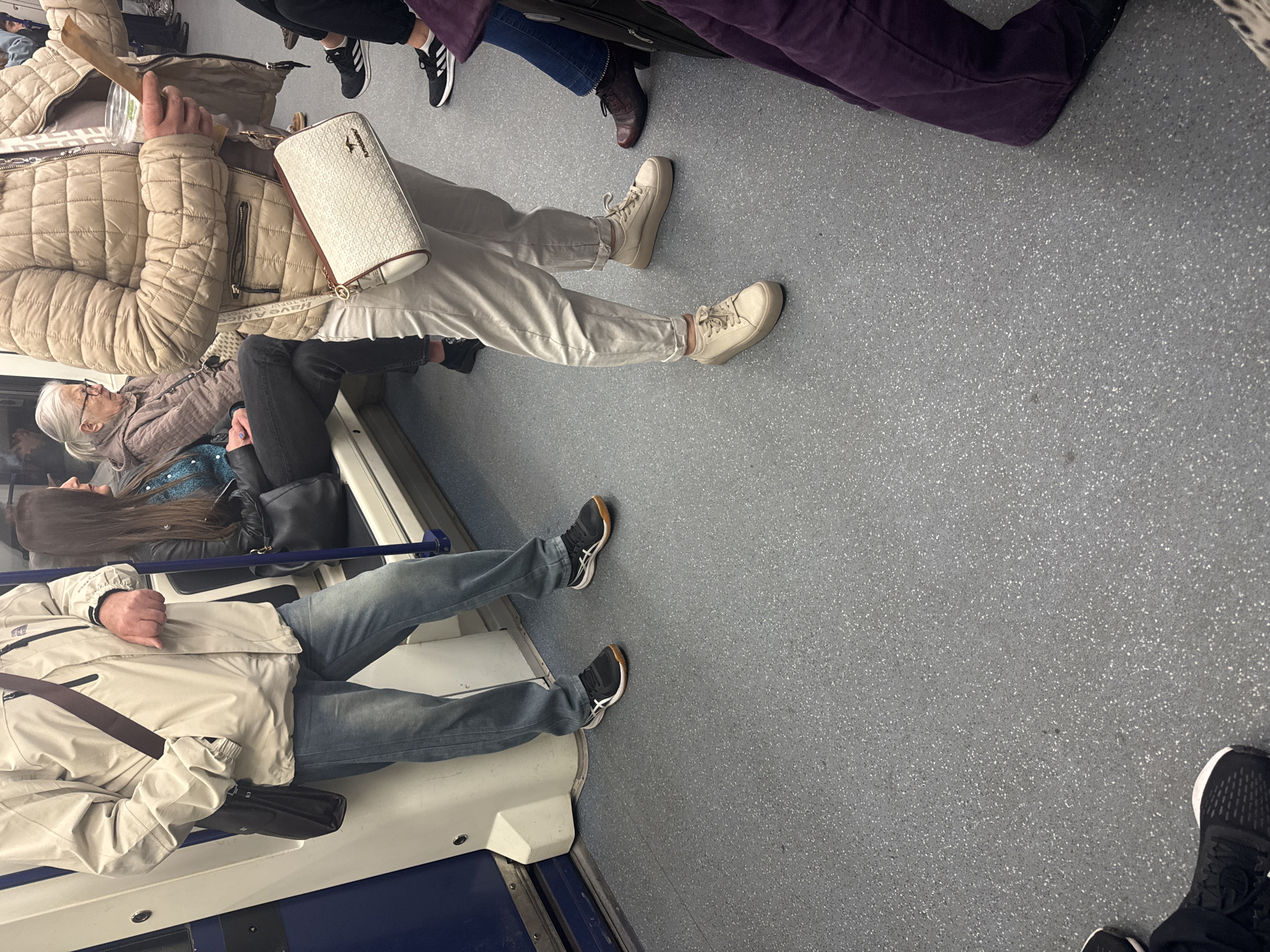 The image shows the interior of a train car, possibly a subway or metro train, with several people seated and standing. The car has a gray floor. The seats appear to be blue and white.
One person is wearing a beige puffer jacket and light-colored pants and has a white purse. Another person is wearing a light-colored jacket and blue jeans. The people are passengers on the train.
The image may be taken in Sofia, Bulgaria, although there is no direct indication that points specifically to the city. The picture looks like the underground train.