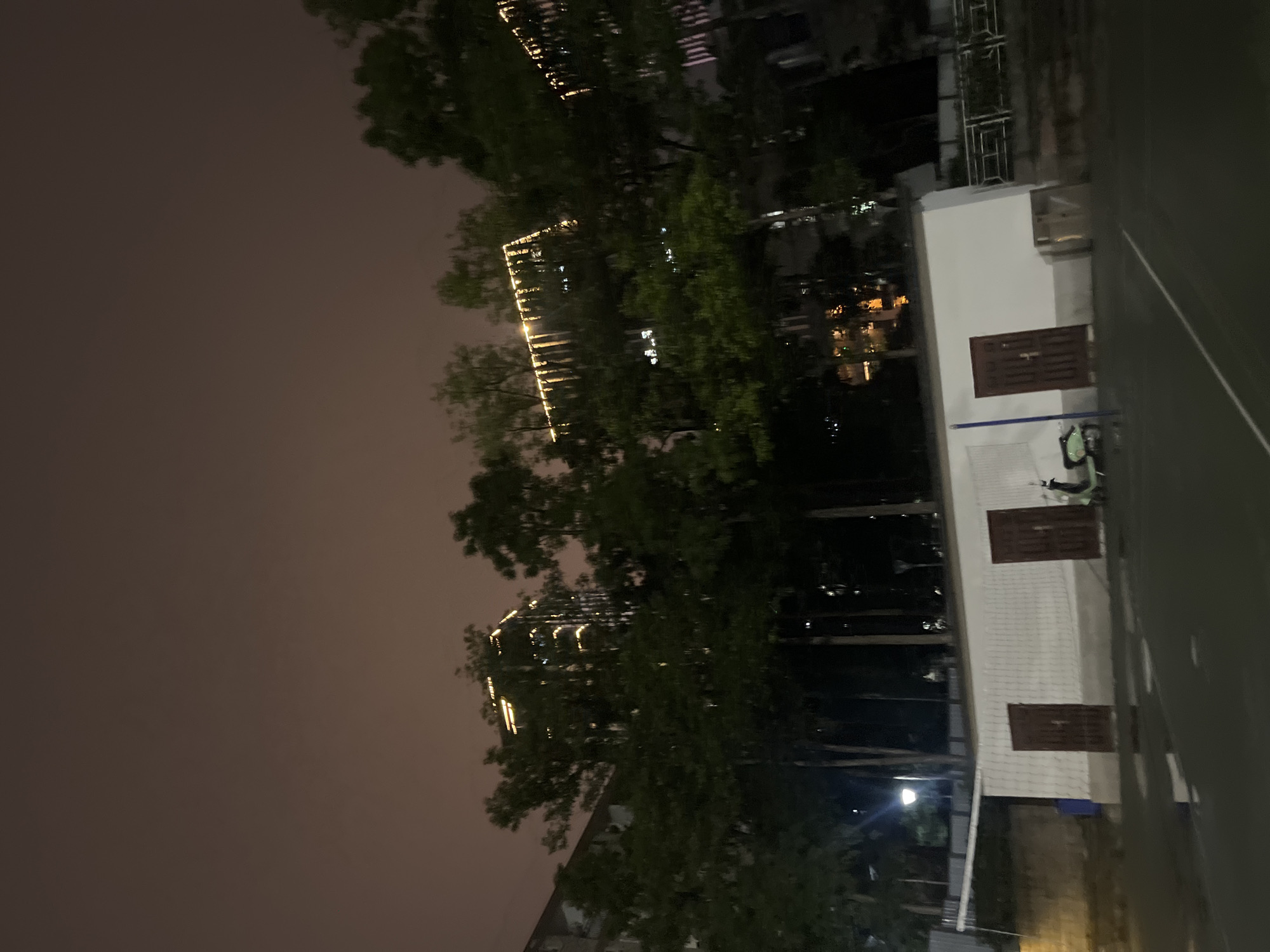 The image depicts a nighttime scene in Yuzhong District, China. The photograph is oriented vertically, capturing a view that includes trees, buildings, and an outdoor area. Tall trees with dark green foliage dominate the foreground and midground, their leaves obscuring parts of the background. In the distance, illuminated by artificial lights, are several tall buildings. These buildings appear to be modern, with windows and decorative lighting. One building, in particular, stands out with horizontal bands of warm yellow lights.
To the right of the image is a low-rise building with a white facade. This building has two dark brown doors, set within recessed panels. Above these doors, there are some architectural details, possibly vents or small windows. A green scooter is parked in front of the building. The ground area in front of this building appears to be a paved surface, possibly a court or a pathway, with white lines visible, suggesting a sports court. Further to the right, a darker, vertical element, likely a wall or another structure, runs along the edge of the frame.
The sky is a muted, hazy gray, indicating a possibly overcast night or the presence of atmospheric pollution. The overall mood is serene and somewhat mysterious, amplified by the nighttime setting and the limited visibility. The lighting is primarily artificial, coming from the buildings and streetlights, casting a warm glow on some elements and deep shadows on others. There is no visible text in the image. The scene suggests an urban environment at night, with a blend of natural elements (trees) and constructed infrastructure.