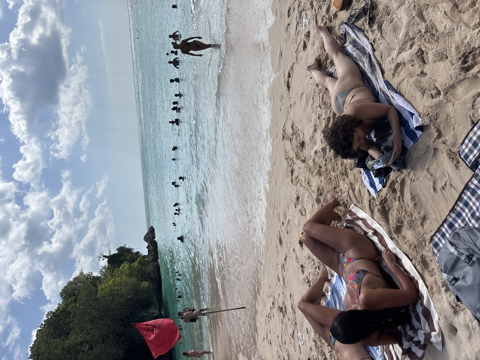 The image depicts a crowded tropical beach scene under a clear, partially cloudy sky, consistent with a location such as Kuta Selatan, Indonesia.
In the foreground, two women are sunbathing on light-colored sand. The woman on the left lies prone on a blue and white striped towel, facing away from the camera, revealing her dark hair and a multi-colored geometric patterned bikini. Her legs are bent at the knees. The woman on the right lies prone on a similar blue and white striped towel, facing left, with her curly dark hair visible. She wears a grey bikini and is actively looking down at a mobile device held in her hands. A silver can is near her left hand, and a brown coconut shell is positioned to her right. A grey duffel bag rests on a plaid blanket in the bottom center. Scattered pieces of white plastic litter are visible on the sand near the water line.
The midground features numerous people wading or swimming in the clear, light blue-green ocean water. A red flag, bearing a dark circular symbol, is planted on a pole in the sand at the left side of the frame, near the water's edge. A woman in a dark bikini walks out of the water towards the sand on the right side of the midground. Another woman in a pink bikini wades near the shore on the far left. Several individuals are visible with their torsos and heads above the water's surface, indicating varying depths.
In the background, the ocean extends to the horizon under a bright sky with scattered white cumulus clouds. Sunlight glints off the water's surface in the distance. A steep, lush green cliff covered in dense vegetation dominates the left background, with rocky outcrops visible at its base extending into the water. The overall lighting suggests midday.