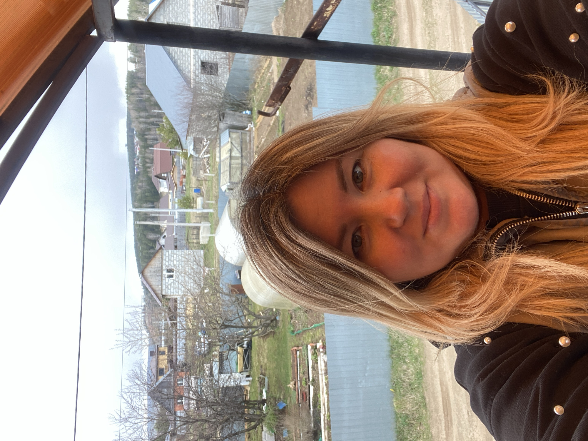 The image is a selfie of a woman taken against a rural landscape, appearing rotated 90 degrees clockwise. When viewed correctly, the woman's face and upper torso are in the foreground, with the background depicting a village scene.
The woman, with long, wavy blonde and light brown hair, is looking directly at the camera with a gentle smile. Her face is well-lit, and her right eye is clearly visible. She is wearing a dark, possibly black, jacket adorned with silver studs or buttons on the shoulder/sleeve, and a zipper is visible on the front.
A dark metal railing or bar runs horizontally across the middle of the image, separating the woman from the distant background. To the top left, a partial view of a translucent orange canopy or structure is visible.
The background shows a village or rural settlement, consistent with the provided context of "Ольховка, Russia." The weather appears to be overcast, with a bright, cloudy sky. Several houses of different constructions are scattered, including one prominently built with concrete blocks and another with a reddish-brown roof. Bare trees, indicating late winter or early spring, are visible throughout the landscape, interspersed with patches of greening grass and dirt paths. A forested hill or mountain range is visible in the distant background. Utility poles and lines, along with what appear to be greenhouses or agricultural structures, add to the rural character of the scene.
The overall mood is calm and pleasant, with the woman appearing relaxed in her environment. There is no visible text in the image.