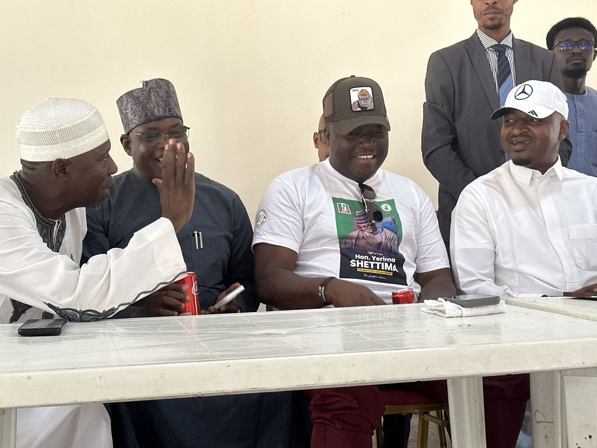 Four men are seated at a white, rectangular table. The man on the left wears a white kufi and matching top. The second man from the left has on a dark blue robe and a matching patterned hat. He is holding up his hand. The third man from the left is wearing a white t-shirt with a graphic of “Hon. Yerima Shettima for Senate 2017 Kaduna Central" on the front, and a dark cap with a cartoon face on it. The man on the right is wearing a white button-down shirt and a white baseball cap with a logo on it. Behind them are two men standing. The man on the left is wearing a gray suit, white shirt, and striped tie. The man on the right is wearing a dark blue shirt and eyeglasses. There are Coca-Cola cans, pens and cell phones on the table. The scene appears to be indoors.