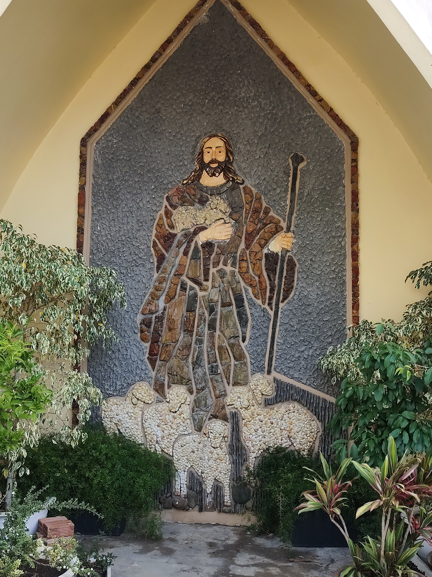 A mosaic depicting Jesus as the Good Shepherd stands in front of greenery. The mosaic is within an alcove with a triangular apex at the top. Jesus, with long brown hair and a beard, is in the center, holding a staff. He wears a robe assembled from different shades of brown. Below him are several sheep made of white stone. The mosaic's background is gray, surrounded by a tan-colored alcove. The base is paved with gray stone. Plants surround the mosaic. This location is in Da Lat, Vietnam.