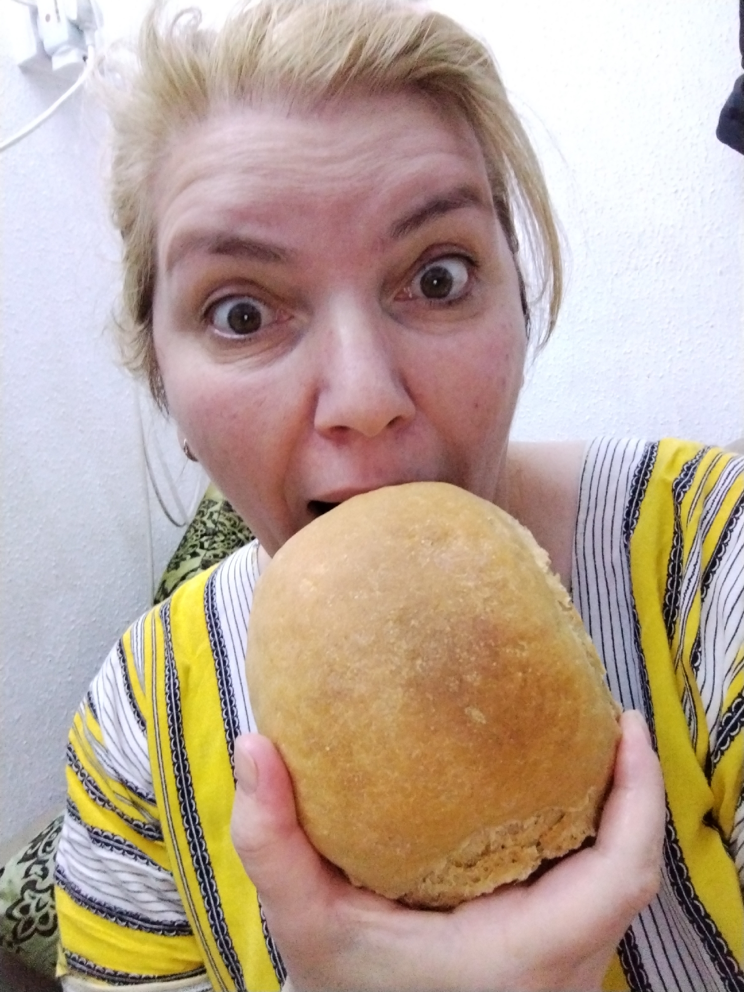A woman with light skin and blonde hair holds a round, light brown roll in her hand, appearing ready to take a bite. She wears a yellow and white striped shirt and looks directly at the camera with wide eyes. The background consists of a textured white wall, with an electrical outlet visible in the upper left corner. Part of a patterned cushion is visible to the left. The image appears to be taken indoors, likely in a home or apartment in the city of Al Mas'oudi, United Arab Emirates.