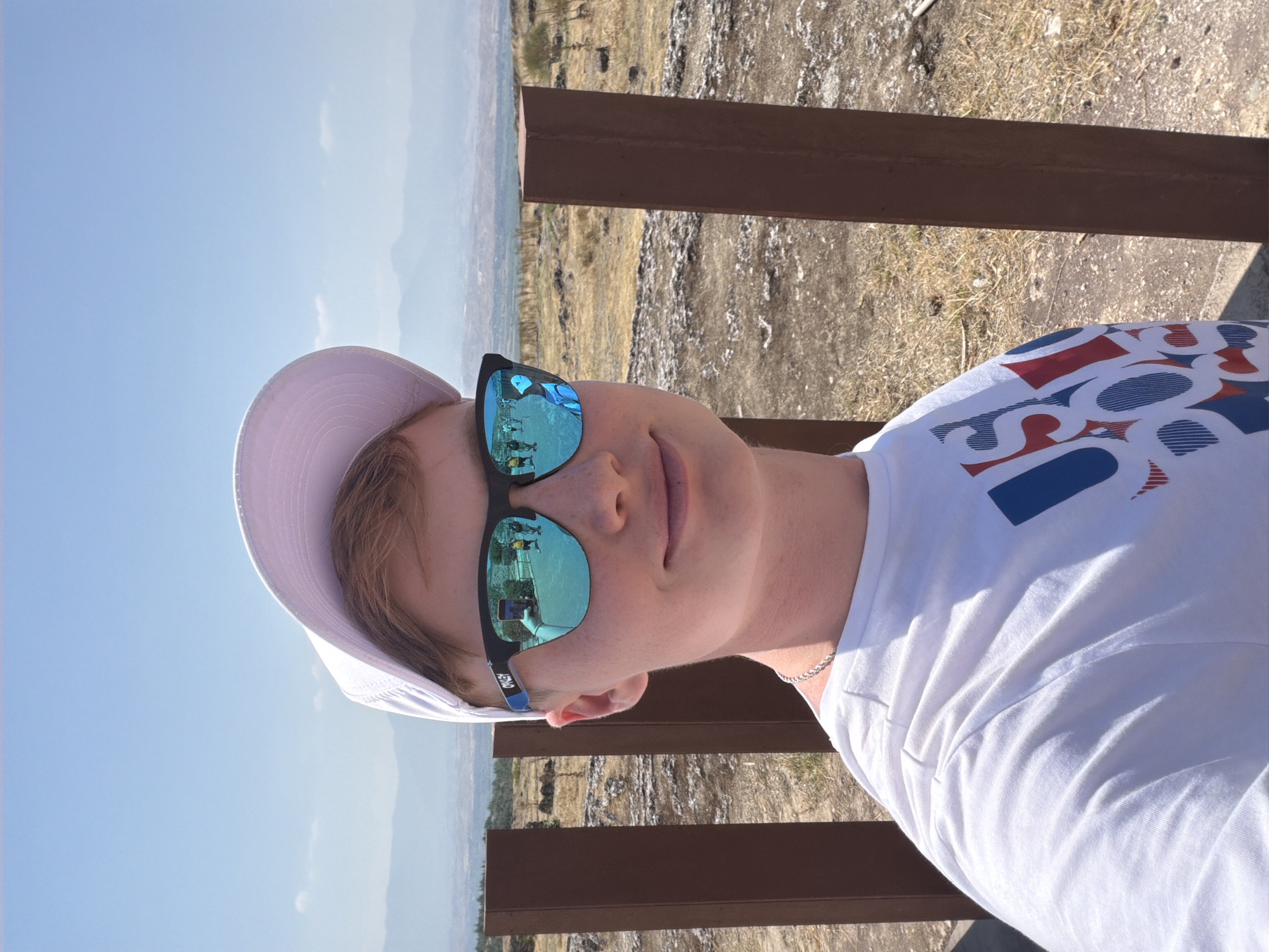 A young person with fair skin, light brown hair, and freckles is taking a selfie outdoors on a sunny day. They are wearing a white baseball cap, mirrored sunglasses, and a white t-shirt with a red, white, and blue graphic design. The person is smiling and looking at the camera. In the reflection of their sunglasses, another person is visible holding a phone, taking a picture. Behind the person, there is a landscape with dry, yellow grass, scattered rocks, and two tall, brown, weathered wooden posts. In the distance, there are hills and mountains under a clear blue sky with a few wispy clouds. This scene appears to be within the ancient city of Hierapolis, located in Pamukkale, Turkey, known for its historical ruins and travertine terraces.