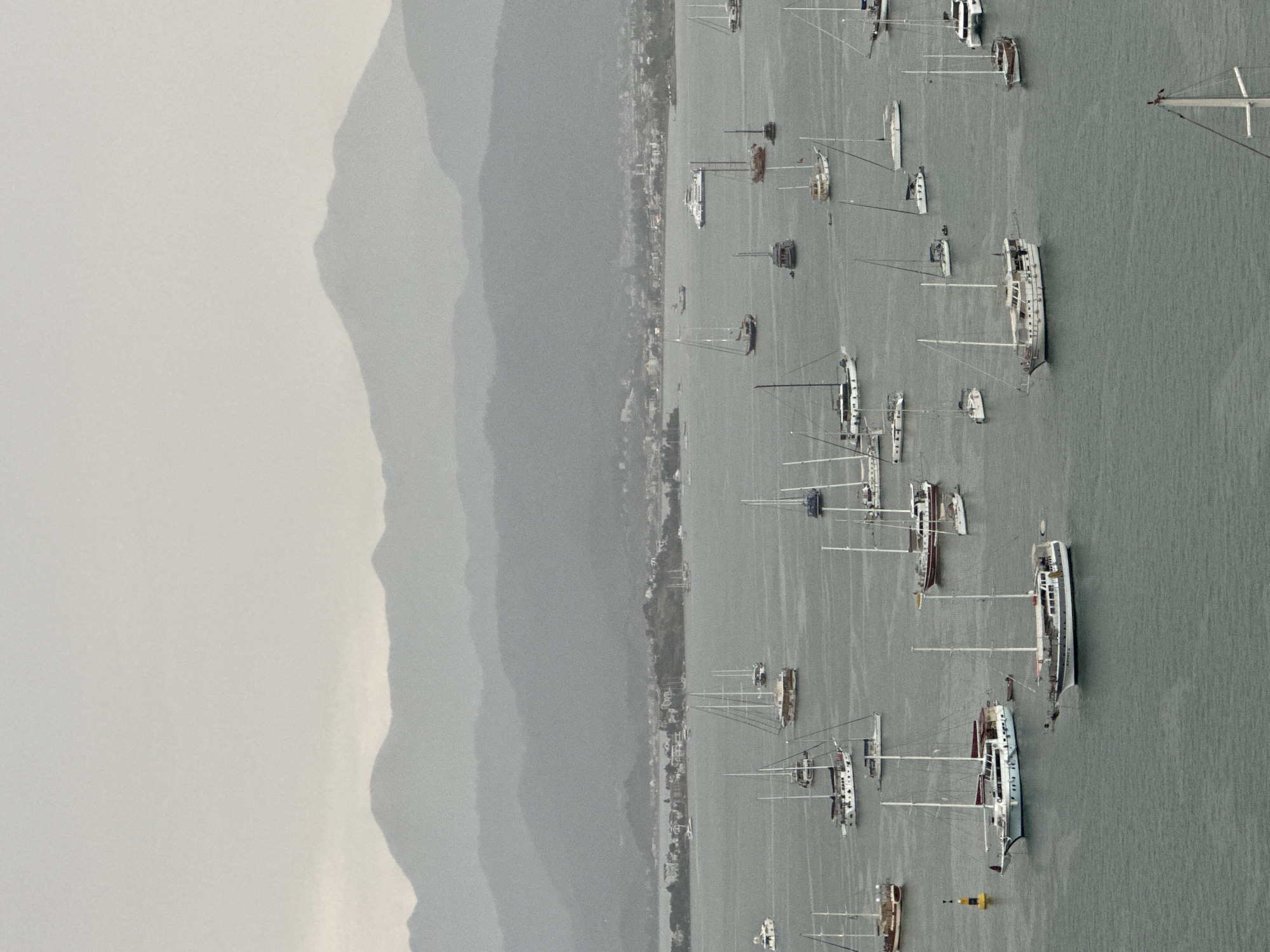 The image shows a panoramic view of Fethiye, Turkey, with a bay filled with numerous sailboats and yachts anchored. The water is calm and grey, reflecting the overcast sky. In the background, a series of layered mountains fade into the distance, shrouded in mist, creating a serene and atmospheric scene. The coastline is visible with buildings and greenery, indicating the presence of the city. The sailboats are of various sizes and designs, with some having sails furled and others with masts visible against the sky. The overall impression is a tranquil harbor scene on a cloudy day in a picturesque coastal city.