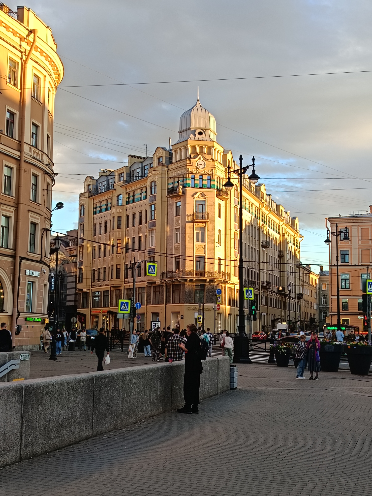 This is a medium-shot, eye-level, outdoor photograph of a street corner in Saint Petersburg, Russia, taken during the golden hour. The light of the setting sun illuminates the facades of the buildings, casting long shadows. In the foreground, a gray concrete wall lines a pedestrian walkway paved with dark cobblestones. A man dressed in black stands with his back to the camera, facing the street. Behind him, a busy street is visible with pedestrians walking and cars driving.
Dominating the center of the image is a large, ornate building with a distinctive domed roof and a clock face on its facade. The building is bathed in warm sunlight. To its left, another building with a curved facade and multiple windows is partially visible. Several street lamps with intricate designs are positioned along the street. Pedestrian crossing signs with yellow diamond shapes are present at various intersections. A traffic light is also visible, currently showing green. The sky above is partly cloudy, with patches of blue sky visible between the clouds, reflecting the warm tones of the sunset. The overall atmosphere is urban and lively.