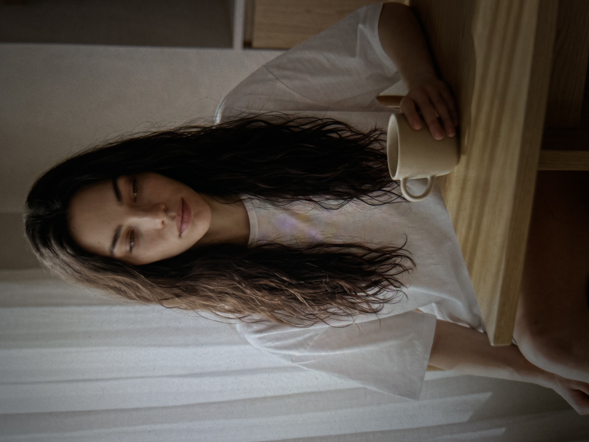 A medium shot shows a woman with long, dark, wavy hair sitting at a wooden table. She is wearing a white t-shirt with a subtle, multi-colored graphic on the left side of her chest. Her left arm is resting on the table, and her left hand is holding a light brown mug with a handle. Her gaze is directed downwards, slightly to her left. The background consists of a sheer white curtain on the left and a neutral-colored wall on the right. The lighting creates soft shadows on the table. The scene appears to be indoors, in a room with natural light coming from a window.