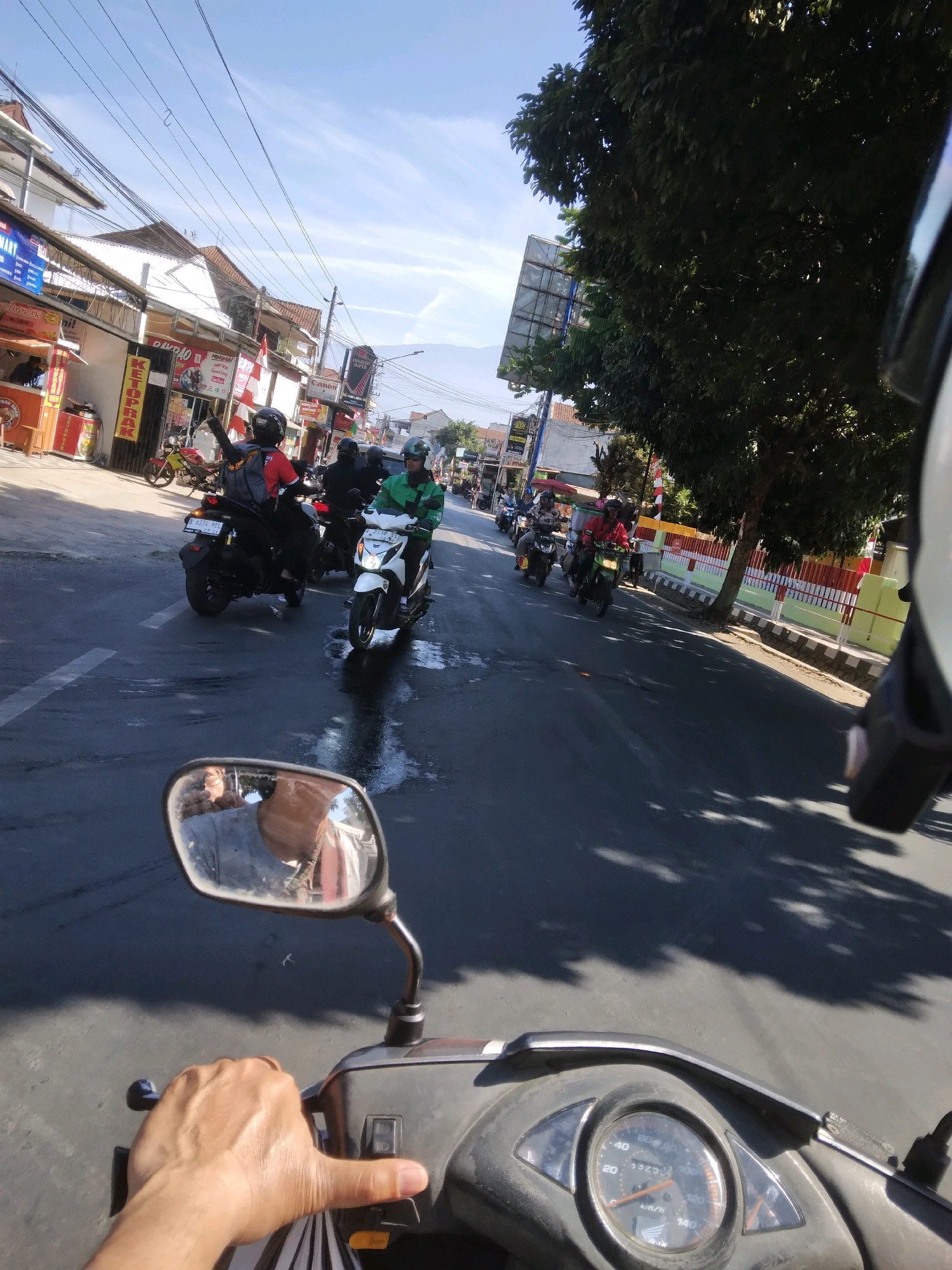 The image shows a first-person perspective of someone riding a motorcycle on a street in Grendeng, Indonesia. The road is wet and reflective, with visible tire marks. Several other motorcycles, ridden by individuals wearing helmets, are also present on the road. Some of the riders are wearing distinct green jackets. On the left side of the street, there are buildings with shops and signage, including a banner that says "KETOPRAK." A large tree with dense foliage is on the right side of the street, casting shadows on the road. The sky is clear and blue, with a few wispy clouds. The motorcycle's rearview mirror is visible in the foreground, reflecting the rider's arm and part of their torso. The motorcycle's dashboard, including the speedometer, is also visible. The overall scene depicts a busy street with ongoing motorcycle traffic.