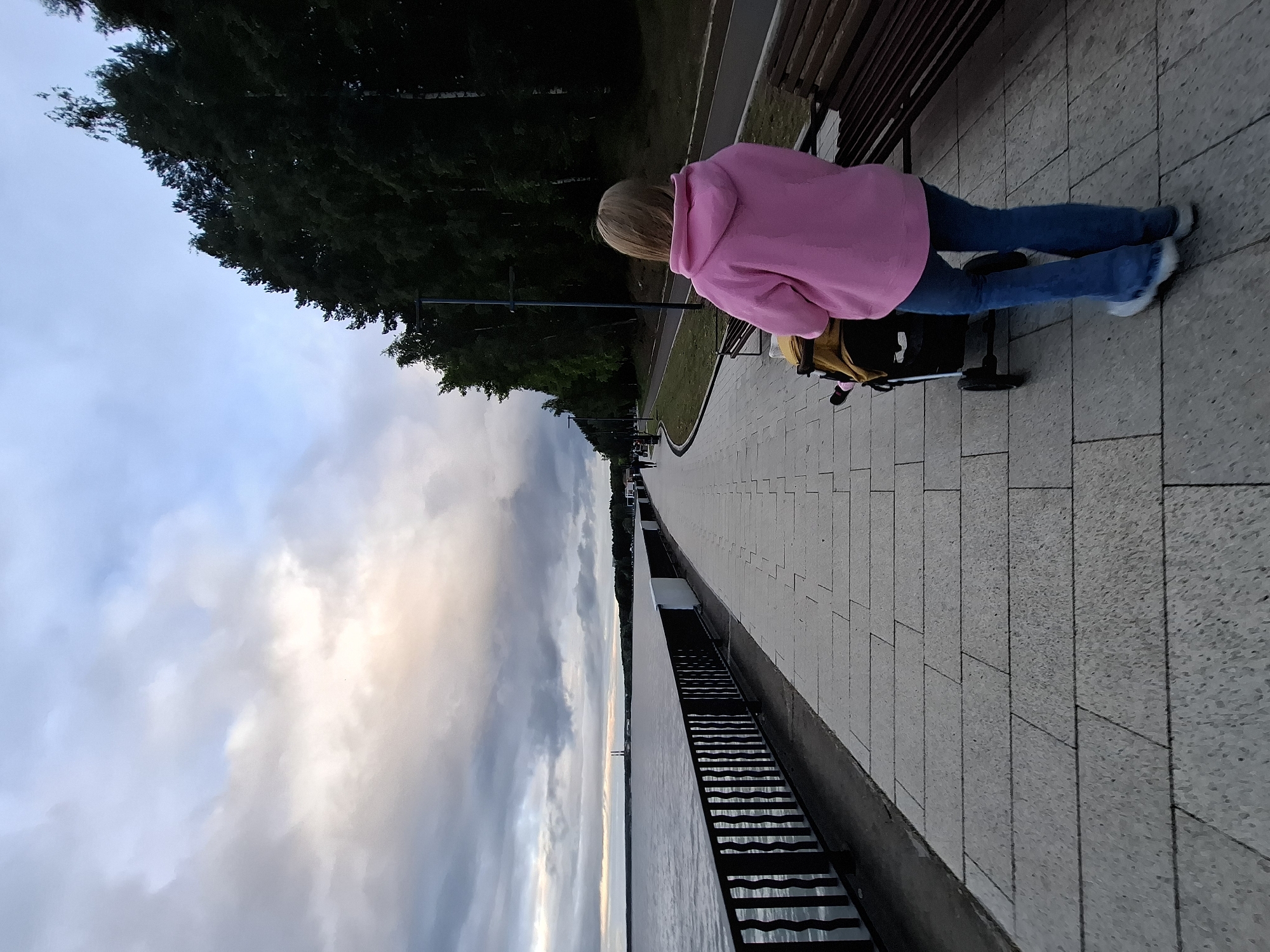 The image depicts a paved waterfront promenade in Chaikovsky, Russia, during dusk under an overcast sky. A single individual, seen from behind, walks away from the viewer, pushing a dark-colored stroller with a yellow upper section. The person is wearing a pink hooded sweatshirt and blue jeans. The promenade is constructed from light grey rectangular paving slabs and extends linearly into the distance. To the left, a black metal railing separates the walkway from a large body of water, exhibiting subtle surface ripples. On the distant horizon across the water, a treeline is visible, along with two prominent vertical industrial structures resembling smokestacks or towers. To the right of the main promenade, a narrower parallel paved path runs alongside a grassy strip, backed by a dense line of dark green trees. Several dark-colored park benches are placed at intervals along the right edge of the promenade. Streetlights are visible within the treeline, indicating the approaching night. The sky displays a mix of grey and blue clouds with a faint warm glow on the horizon.