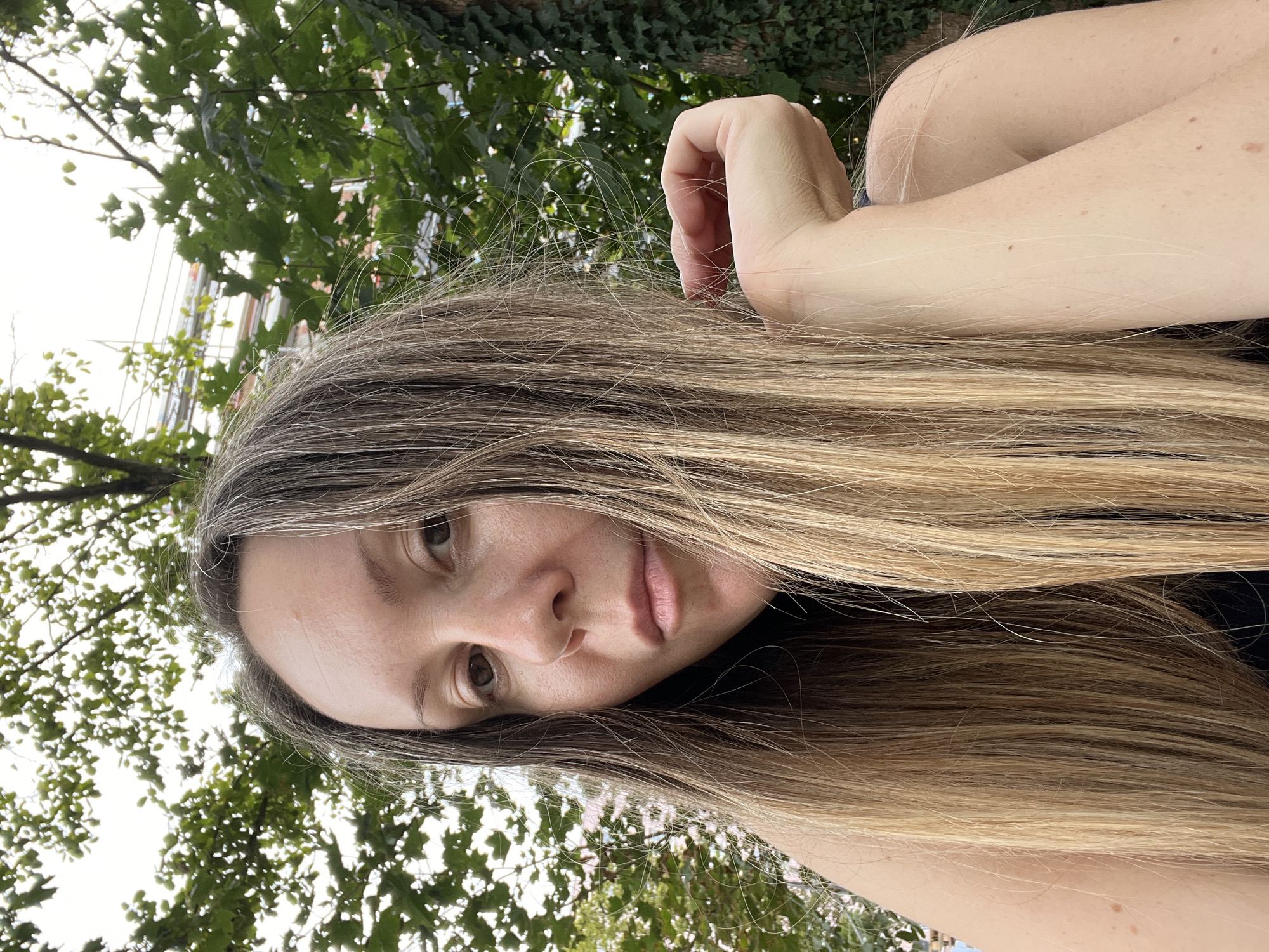 A woman with long, blonde hair and brown eyes is looking towards the camera. Her head is tilted slightly to the left, and her right hand is resting on her chin, with her fingers extended. She is wearing a dark-colored top. The background is out of focus and shows green foliage and trees, suggesting an outdoor setting. The image is a close-up of the woman's face and upper body. The location of the image is not discernible from the visual information provided.