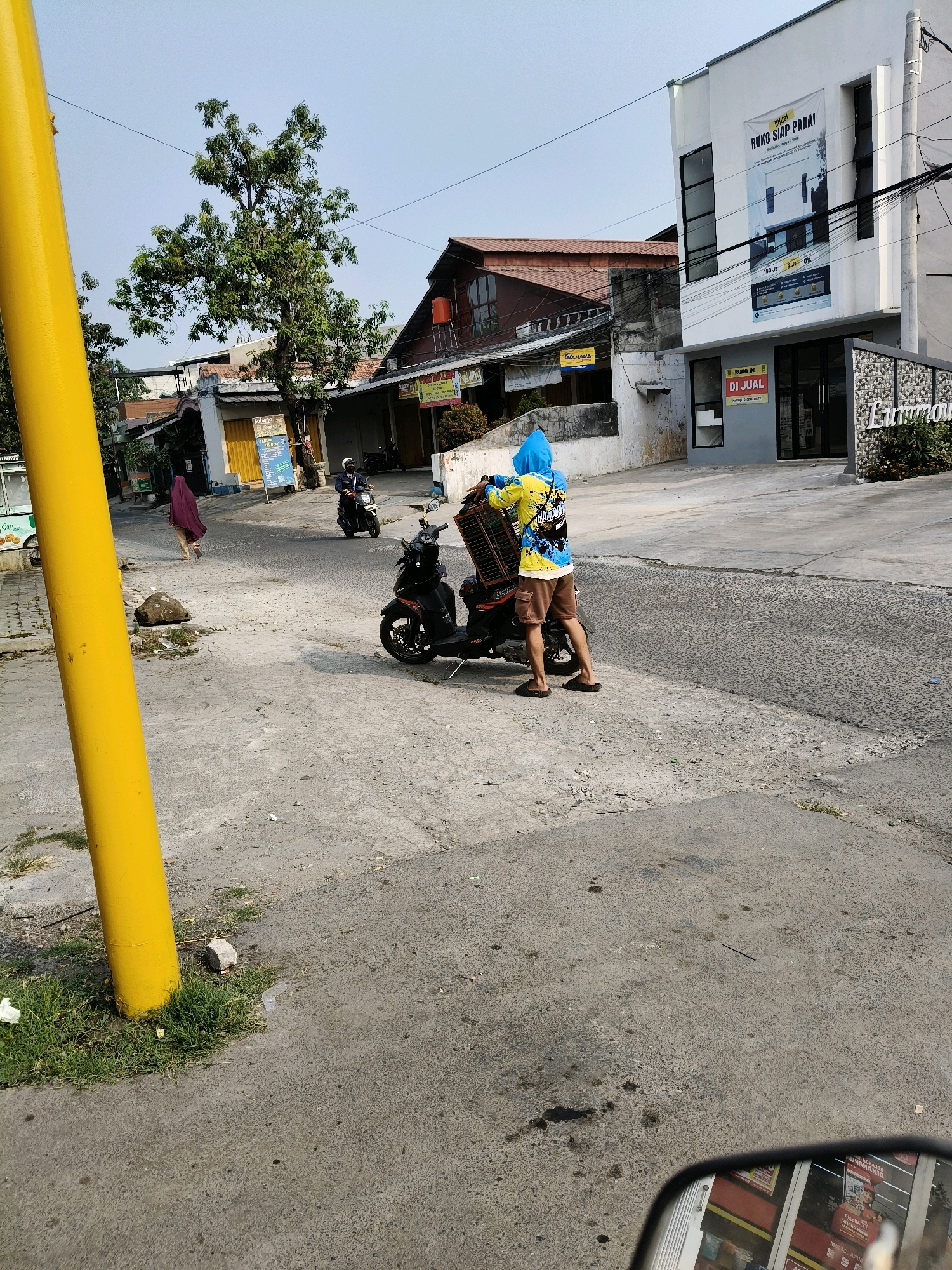 The image depicts a street scene in Setiamekar, Indonesia. A person wearing a bright blue hooded sweatshirt and brown shorts is standing next to a black motorcycle. The person is holding a wicker basket on top of the motorcycle. The motorcycle is parked on the side of a paved road. To the right of the motorcycle is a modern white building with dark windows. A sign on the building reads "DI JUAL RUHO SIAP PAKAI". Further to the right, a sign with decorative metalwork reads "Luminous". In the background, there are other buildings and trees. A yellow pole is visible in the foreground on the left side of the image. The sky is clear and bright.