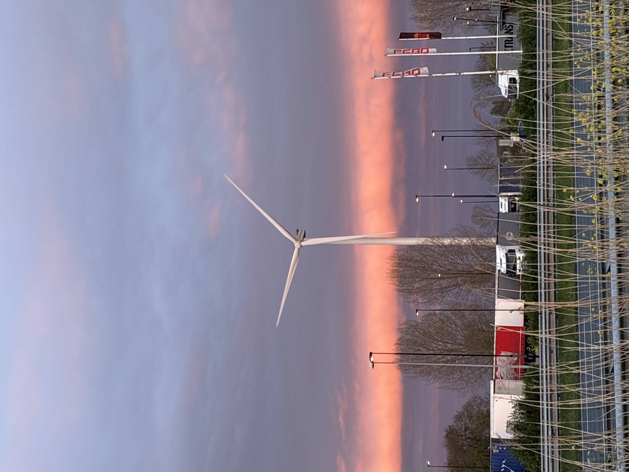 A vertically oriented photograph captures a scenic view in Bruchem, Netherlands, showcasing a wind turbine against a twilight sky. The sky is divided into a grayish-purple hue on the left and a band of pinkish-orange to the right. A wind turbine, positioned toward the left side of the frame, reaches into the sky. To the right of the wind turbine, along the ground, is a row of street lights, followed by a row of commercial vehicles. Beyond the vehicles, there is a metallic fence lined with vegetation. The flags on the right appear to show the word FEBO. Overall, the photograph combines industrial elements with the natural beauty of a sunset.