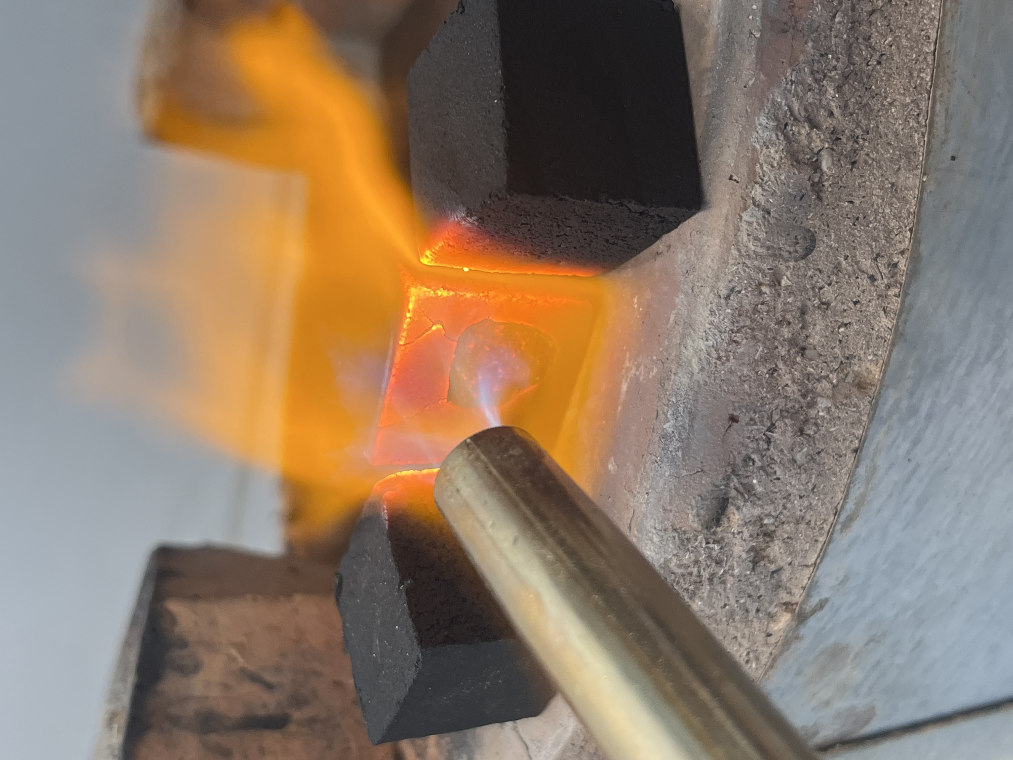 A close-up shot depicts a gold-colored blowtorch applying heat to a metal container, likely a crucible, nestled between four dark, cube-shaped objects, possibly charcoal briquettes. The flame from the torch is orange and intensely concentrated on the metal within the container, which appears to be glowing. The crucible sits inside a concrete structure that appears aged. The location is Bac Nha Trang Ward, Vietnam.