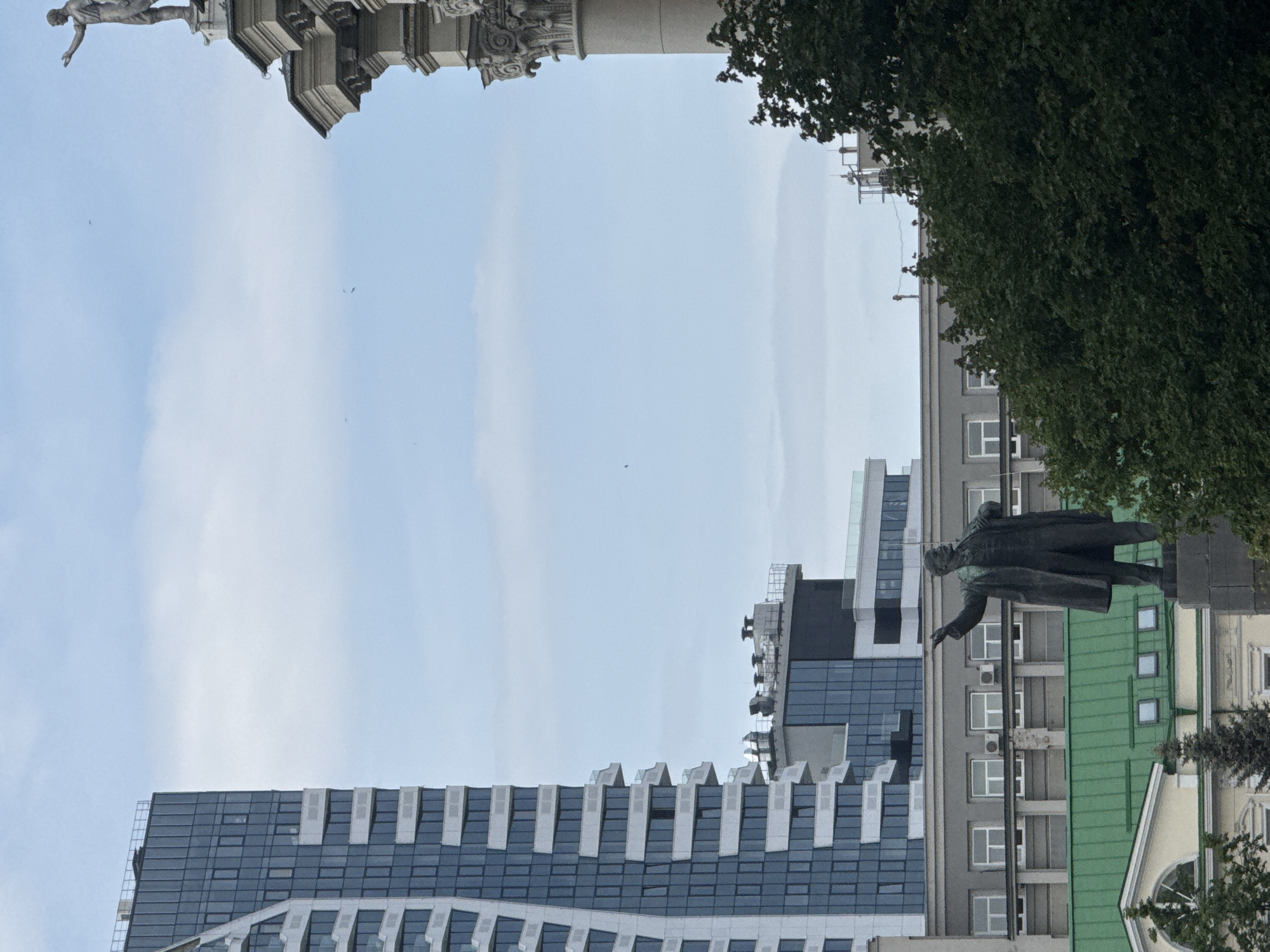 The image shows a view of Yekaterinburg, Russia, featuring architectural elements and landmarks. At the top left, there is a statue atop a column, partially visible. The sky is overcast with light blue and white tones. Below, a modern building with a structured facade of alternating gray and white bands is seen. To the right, a statue of a man in a coat, likely a historical figure, stands in front of a multi-story building with green roofing. Trees are visible to the right, framing the scene.