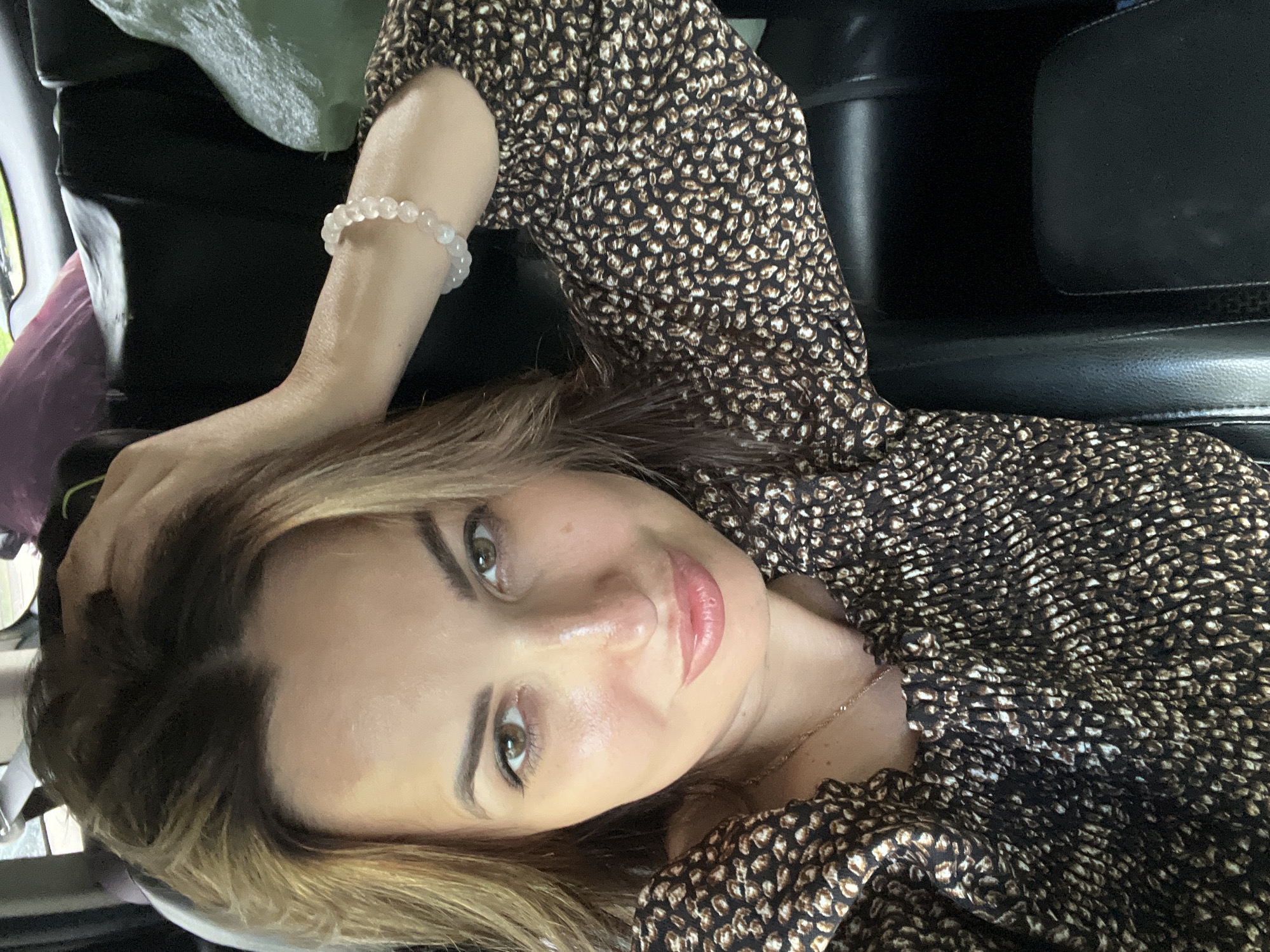 A woman is captured in a close-up, selfie-style photograph, with her head tilted and one hand running through her hair. She has light brown hair, fair skin with a light dusting of freckles across her nose and cheeks, and hazel eyes. She is wearing a dark-colored, patterned top with a smocked neckline and sleeves. A delicate gold necklace is visible around her neck, and she has a bracelet of small, light-colored beads on her left wrist. The background appears to be the interior of a vehicle, with dark upholstery and what seems to be luggage or bags in the upper right corner. The lighting suggests an indoor or possibly overcast outdoor setting.