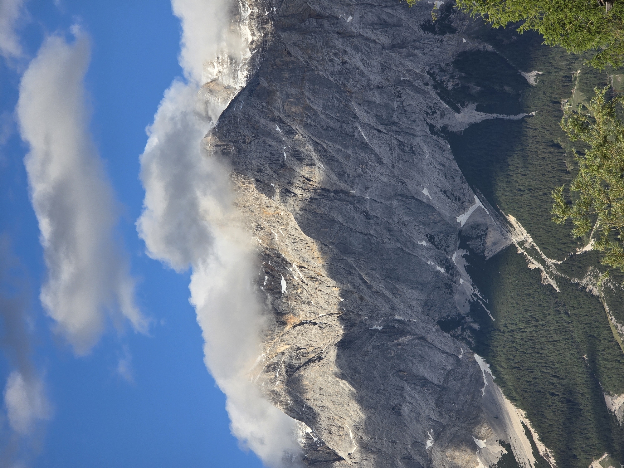 The vertically oriented image captures a dramatic Alpine mountain landscape, likely near Lermoos, Austria. On the left, a clear blue sky is visible, adorned with several large, white cumulus clouds, some stretching vertically across the frame.
The central and right portions of the image are dominated by a massive, rugged mountain face. This imposing peak is primarily dark gray, characterized by deeply textured rock, vertical striations, and scattered patches of snow or ice that gleam in the sunlight. Sunlight illuminates specific sections of the mountain, creating strong contrasts with shadowed areas and highlighting the rugged geology. Clouds cling dramatically to the mountain's flanks and summit, partially obscuring its highest points and creating an atmospheric, mystical feel. Some lighter-colored rock formations are visible through or above the cloud cover.
To the far right, vibrant green foliage from evergreen trees frames the scene, suggesting the viewpoint is from an elevated position, looking across a forested area or a valley. Below the steep rocky face, a dense, dark green forest covers a descending slope into a valley. Within this valley, lighter-colored natural features, possibly scree slopes or exposed rock, meander through the trees.
The scene depicts daytime with strong sunlight and clear shadows. The weather is partly cloudy, with localized cloud formations around the mountain peaks. No people, man-made objects, or visible text are present in the image. The overall impression is one of majestic natural grandeur and isolation.