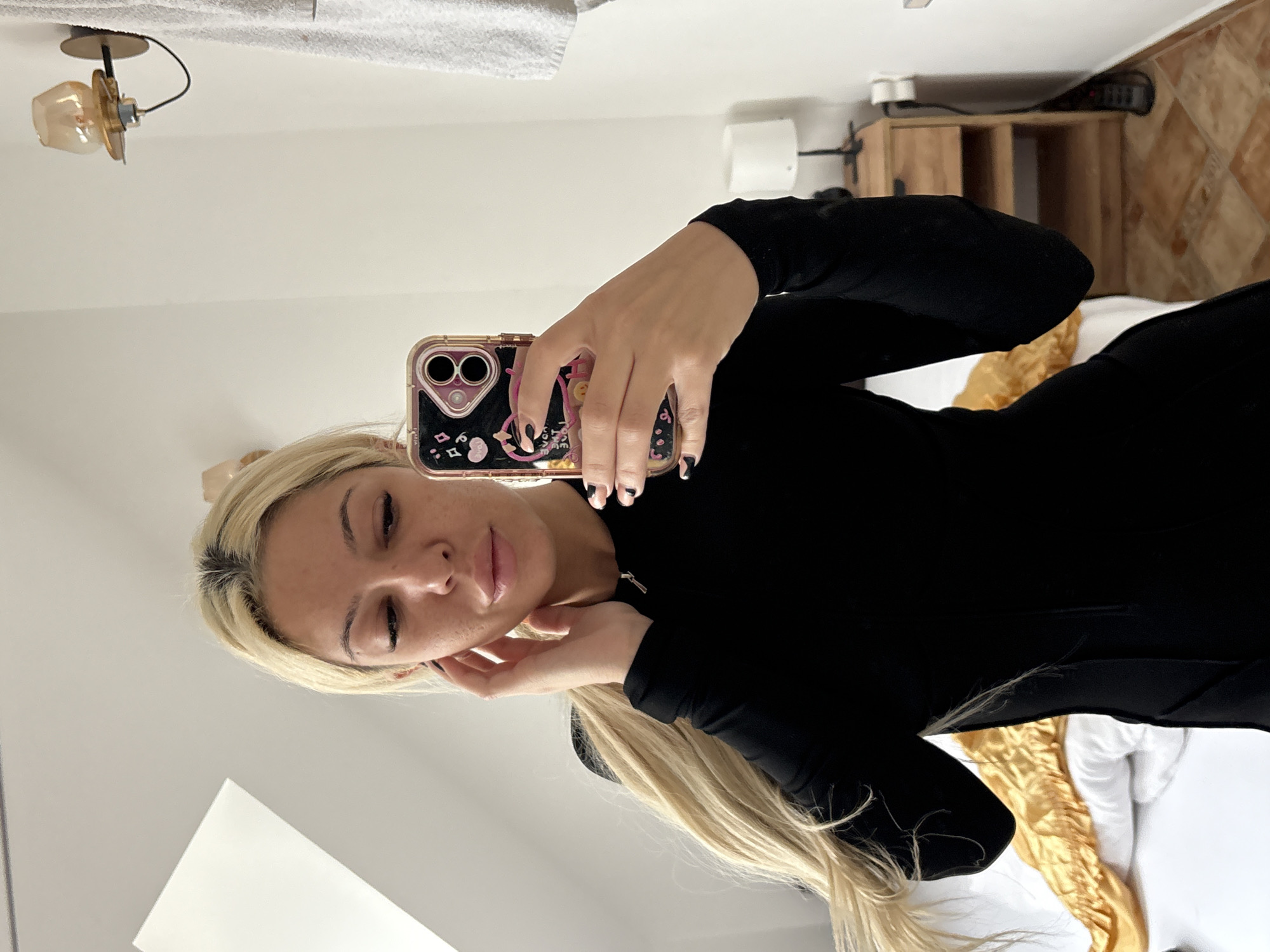 A woman with long, straight blonde hair and visible dark roots takes a mirror selfie in what appears to be an indoor room, likely a bedroom or hotel room. She wears a fitted long-sleeved black top or dress. Her left hand holds a smartphone with a clear rose gold/pink case featuring black and white cartoonish designs; her nails are painted dark with light accents. Her right hand is gently placed near her jawline. Her eyes are closed or looking downwards, and her lips are slightly parted with a natural or light pink gloss.
The background shows clean, white walls. To the upper left, a white textured towel is visible, and above the woman's head, an antique-style wall or ceiling light fixture with a brass base and a warm-toned glass shade is mounted. To the right, a wooden nightstand or small cabinet with open shelves can be seen. Below this, a tiled floor with brown and tan geometric patterns adds a traditional element. A bed with white and golden-yellow bedding or fabric is visible in the lower right portion of the image. The setting suggests a modern European interior, fitting for a location like Sofia, Bulgaria. The overall mood is calm and posed. No explicit text is clearly legible in the image, aside from decorative elements on the phone case.