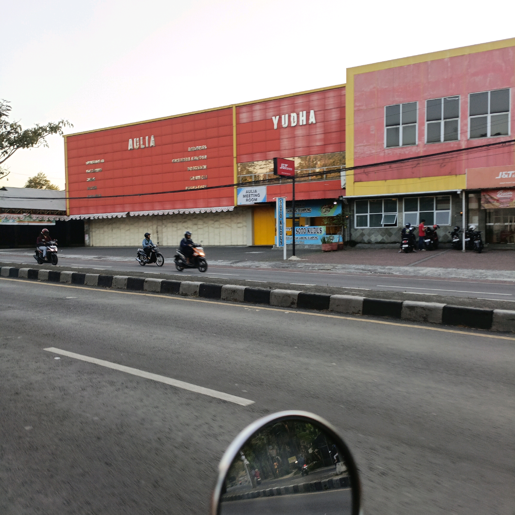 The image shows a street scene in Salatiga, Indonesia, featuring a row of buildings with prominent signage. The buildings are painted in bright colors, primarily red and yellow, and have a modern commercial architectural style. On the red building, large white letters spell out "AGLIA" and "YUDHA". Below these, smaller text lists what appear to be business names or services. A sign for "J&T Express" is visible, as is a blue sign for "AULIA MEETING ROOM" and a vertical sign that says "SOTO KUDUS".
In the foreground, a road is visible with a black and white striped curb. Several people are riding motorcycles on the road. One motorcycle rider in a red shirt and helmet is in the left lane, while another rider in a blue shirt and helmet is further ahead in the same lane. A third motorcycle rider in an orange shirt and dark helmet is in the right lane, overtaking the other two.
In the background, behind the buildings, trees and other structures are visible. The sky is overcast. The overall impression is of a busy commercial area in an Indonesian city, likely during daylight hours.