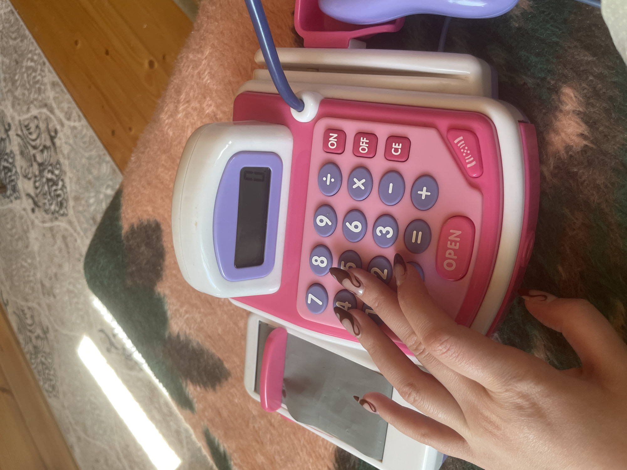 Here is a detailed description of the image:
The image shows a pink and white toy cash register on a patterned blanket or rug. A hand with manicured nails is pressing the number "8" button on the cash register. The cash register has other buttons labeled with numbers and mathematical symbols such as +, -, x, ÷, =, and buttons for "ON," "OFF," "CE," and "OPEN." There is a screen on the left side of the cash register and a drawer below the buttons that is partially open. The background includes a wooden floor and part of a patterned rug or blanket. The location, Zav'yalovo, Russia, cannot be determined from the image content.
