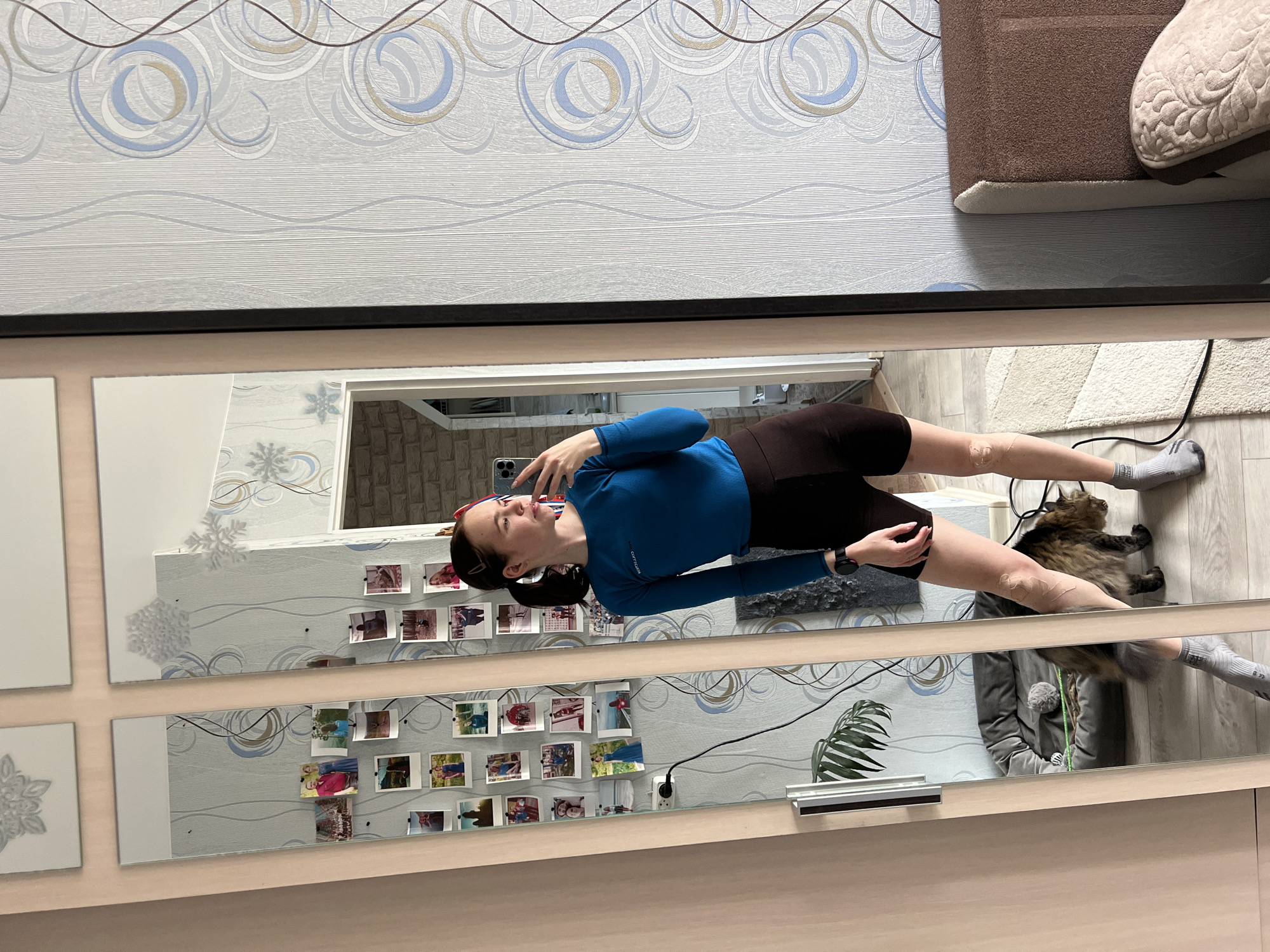 A woman with dark hair pulled back in a ponytail and wearing a blue long-sleeved shirt and dark athletic shorts stands in front of a large mirror, taking a selfie with her smartphone. She is oriented vertically in the frame. A brown and black long-haired cat lies on the floor near her feet. The floor is light-colored wood. A gray upholstered object sits in the upper right corner. The wall visible in the mirror has a patterned wallpaper. Small photographs are attached to the wall reflected in the mirror. A small potted plant is visible in the lower section of the mirror's reflection. The location is Chaikovsky, Russia.