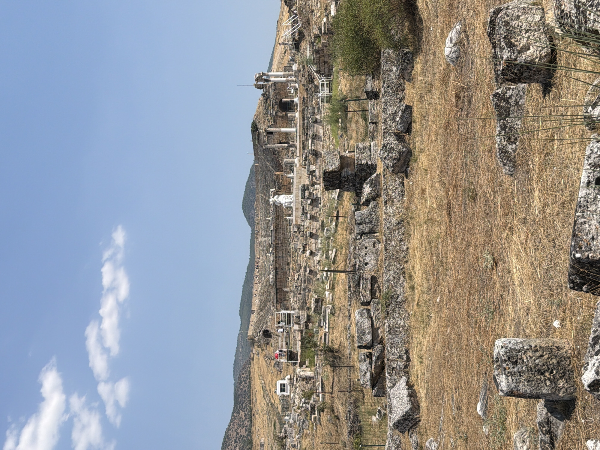 The image depicts the Hierapolis ancient city in Pamukkale, Turkey, under a clear blue sky with scattered white clouds. In the foreground, dry, golden grass covers the ground, interspersed with large, weathered rocks and fallen stone blocks. Some of these stones are rough-hewn, while others appear to be remnants of ancient structures, with some showing signs of erosion.
Further back, the landscape transitions to more ruins. The middle ground is dominated by the remains of a large amphitheater, with tiered seating built into the hillside and sections of stone walls and columns still standing. Several white metal railings and staircases have been erected to guide visitors through the archaeological site. A small white booth or structure with a red umbrella on top is visible to the left, where a few people are standing, appearing to be tourists observing the ruins.
In the background, the rolling hills are sparsely covered with dry vegetation. The overall impression is one of a vast, ancient archaeological site bathed in bright sunlight, showcasing the remnants of a once-grand city. The scene is tranquil and indicative of a historical tourist destination.