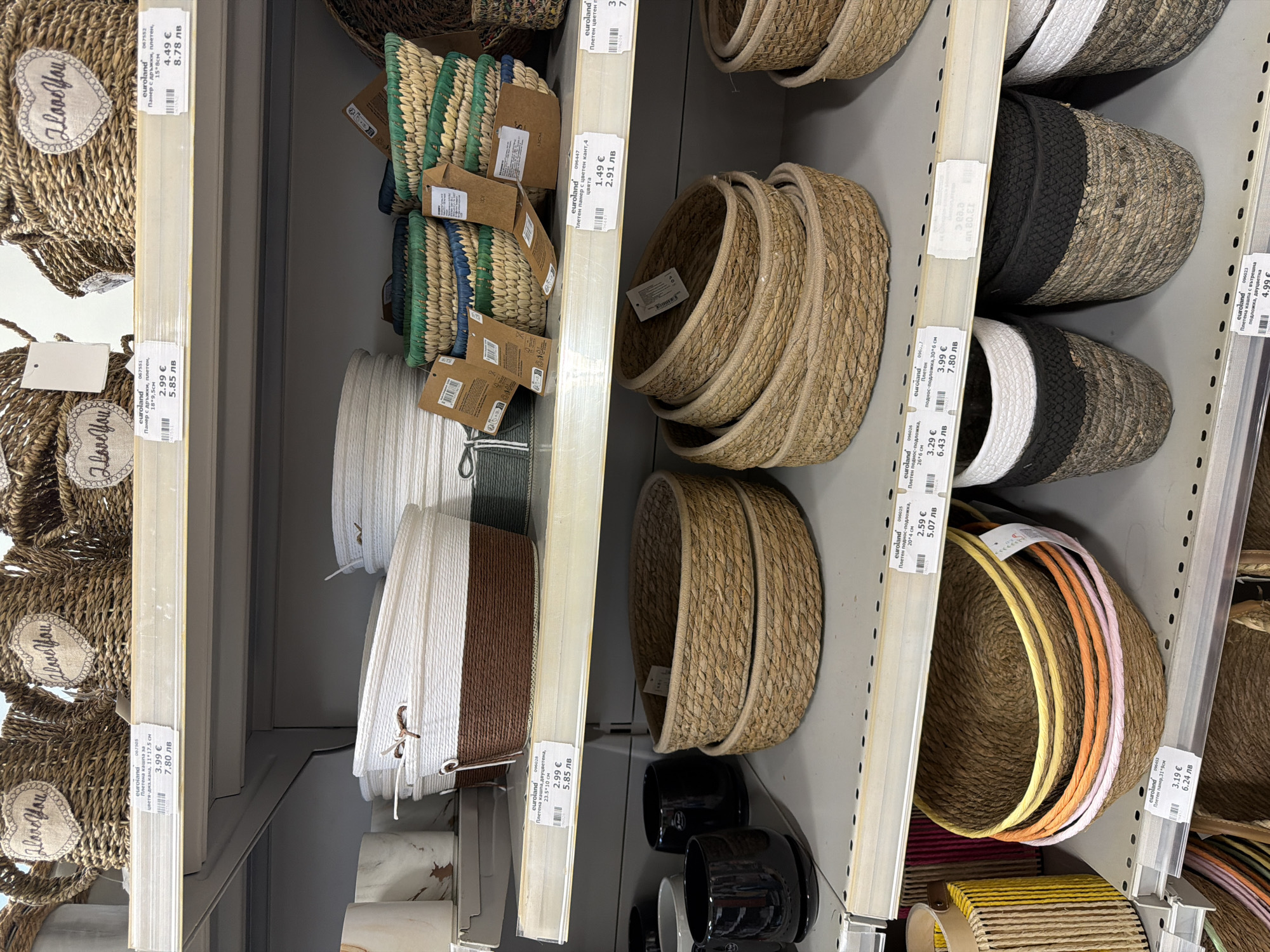 The image shows a retail display of woven baskets in a store. The baskets are made of natural materials and vary in size, shape, and color. Some are plain, while others have decorative elements such as braided handles or colored rims. Several baskets are stacked on shelves, and some have price tags attached. In the foreground, a variety of baskets are visible, including round and oval shapes. The background shows more shelves filled with similar items. The environment appears to be an indoor retail space, likely a home goods or decor store. The lighting is bright and even, suggesting fluorescent or LED lighting common in commercial settings. No people are visible in the image. The context of Pazardzhik, Bulgaria is assumed to be the location of the store.