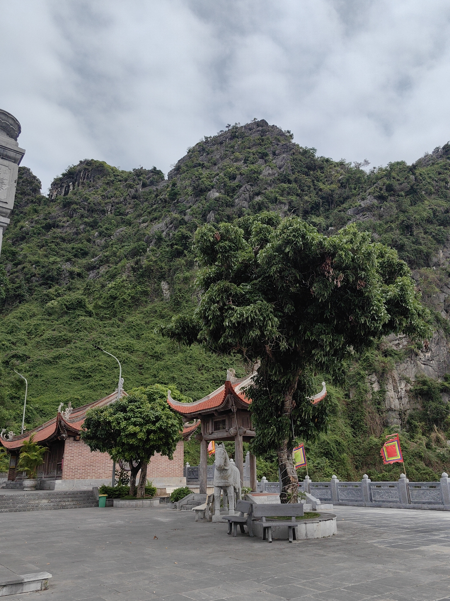 The image depicts a temple complex situated in the city of Hong Gai Ward, Vietnam, against a backdrop of a verdant, mountainous landscape. The foreground features a stone-paved plaza, upon which are positioned benches, a stone horse statue, and a tree with square flags tied around its trunk. A building with a traditional Vietnamese roof and ornate decorations is to the left of the tree. Further back, a lower structure with an ornately decorated Vietnamese roof stands. The background is dominated by a steep, rocky mountain covered with dense vegetation. The sky above is overcast, suggesting a cloudy day. A light post and a grey stone railing with embossed decoration are also visible. No people are apparent in the scene.