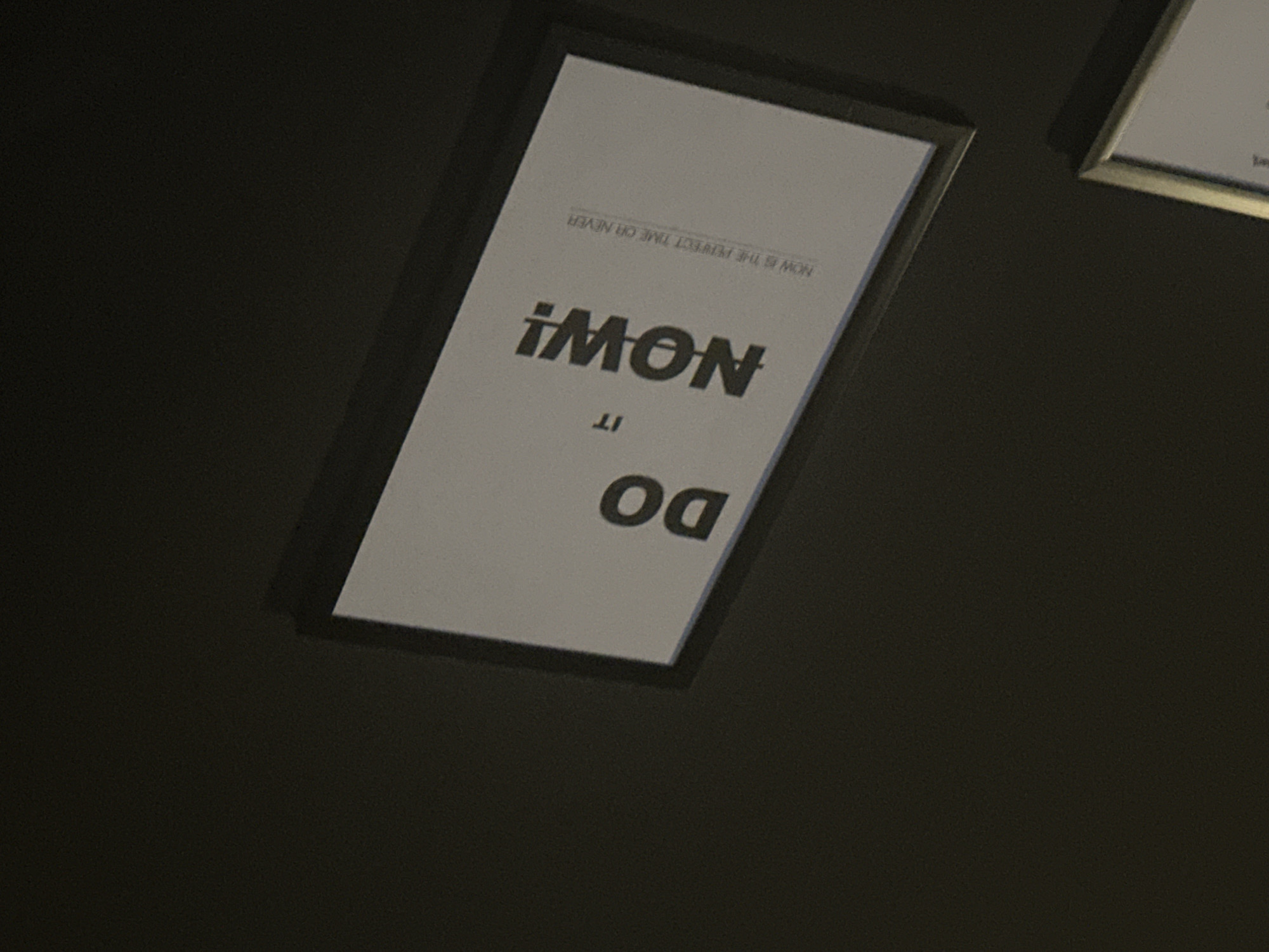 The image captures a close-up, upward-angled view of a framed poster hanging on a dark, possibly black, wall or ceiling. The poster is rectangular with a white background and features dark typography.
The text on the poster is upside down from the viewer's perspective. When mentally rotated to be upright, the text reads:
- "NOW IS THE PERFECT TIME OR NEVER" (smaller font, at the top)
- "DO" (larger font, below the first line)
- "IT" (smaller font, below "DO")
- "NOW!" (largest, bolded font, below "IT", with an exclamation mark).
The frame surrounding the poster appears to be black or very dark. Part of a second framed picture is visible in the top right corner of the image, also with a dark frame and a light interior, suggesting a similar style. The background is uniformly dark, implying an indoor setting with subdued lighting, contributing to the slightly grainy texture of the image. No people or specific events are depicted. The location context provided is Jos, Nigeria, though the image itself offers no explicit visual cues to confirm this.