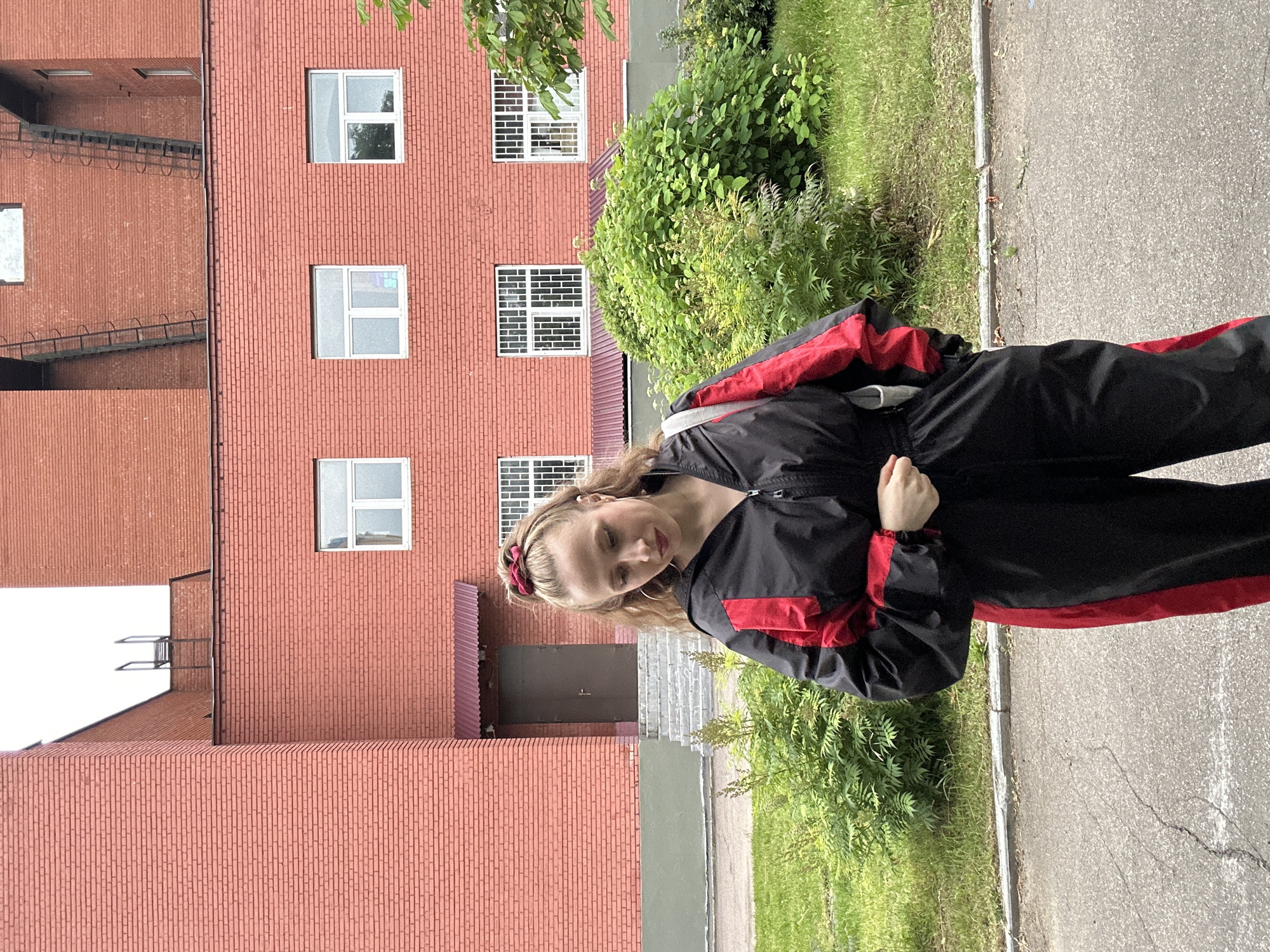 A young woman with blonde hair, wearing a black and red jacket, stands outdoors in front of a red brick building. She is looking down and to her right, with a slight smile. Her hair is pulled back with a red scrunchie, and she is wearing large hoop earrings. The building appears to be a multi-story apartment complex with numerous windows. Green trees and grass are visible to the right of the building, bordering a paved walkway. The scene suggests a daytime setting, possibly late morning or early afternoon, with clear weather. The presence of the building and the greenery suggests an urban or suburban environment in Moscow, Russia. There is no visible text in the image.