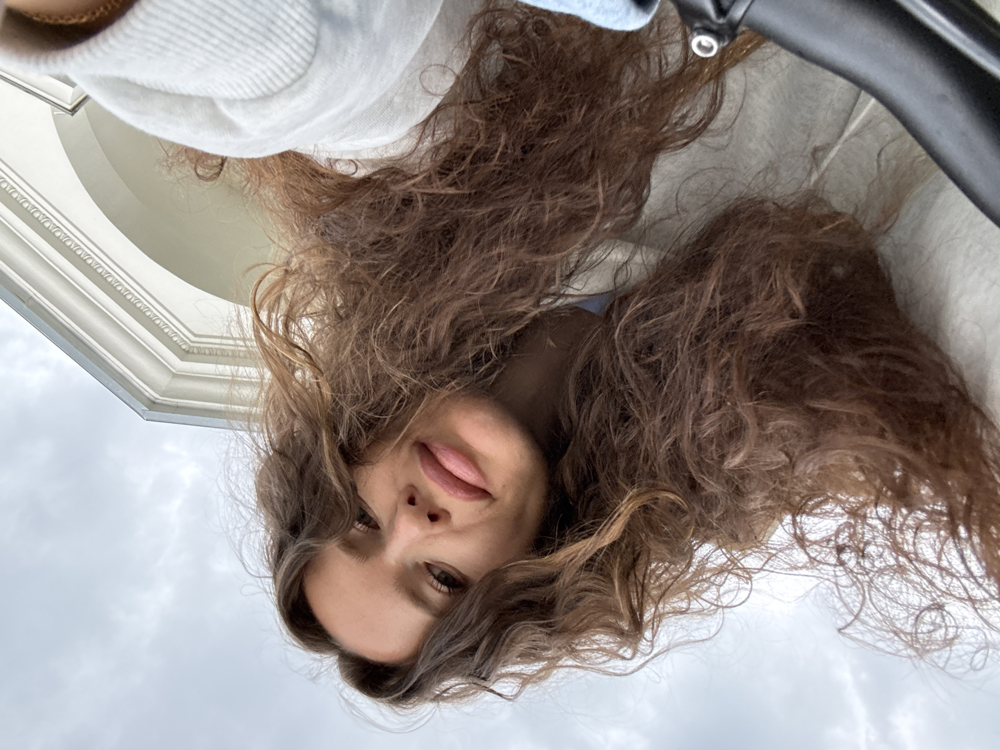 A low-angle, close-up selfie taken outdoors under a cloudy sky. The subject is a woman with long, wavy brown hair, captured from the chest up. She is looking towards the camera with a slight smile. Her facial features include prominent cheekbones and full lips. She is wearing a light-colored hooded jacket. In the background, a portion of a white building with an arched opening and decorative molding is visible. The overall impression is informal and candid. There are no visible indications of the specific location within Moscow, Russia.
